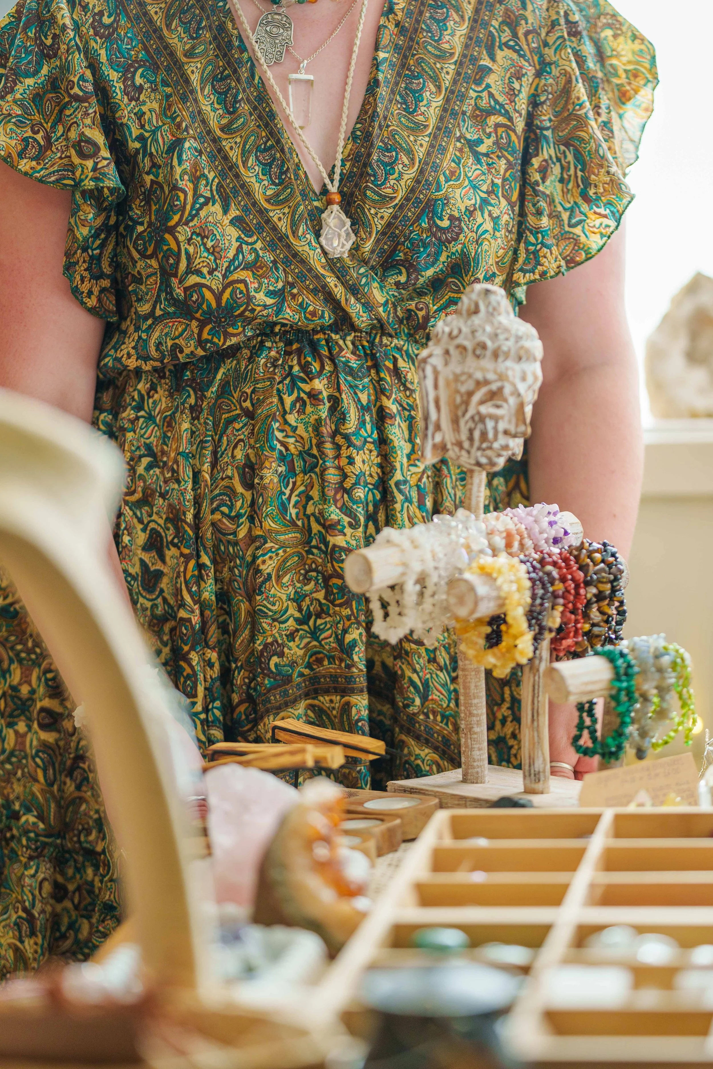 Person wearing a paisley-patterned dress with layered necklaces at a jewelry booth with beaded bracelets and carved Buddha head display.