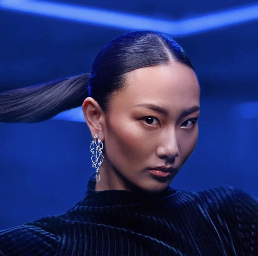 An asian woman with her hair styled in a sleek, low ponytail, wearing statement earrings and a dark, textured top against a blue background. Hair done by Aziza Rasulova for  jewelry photoshoot in NYC.