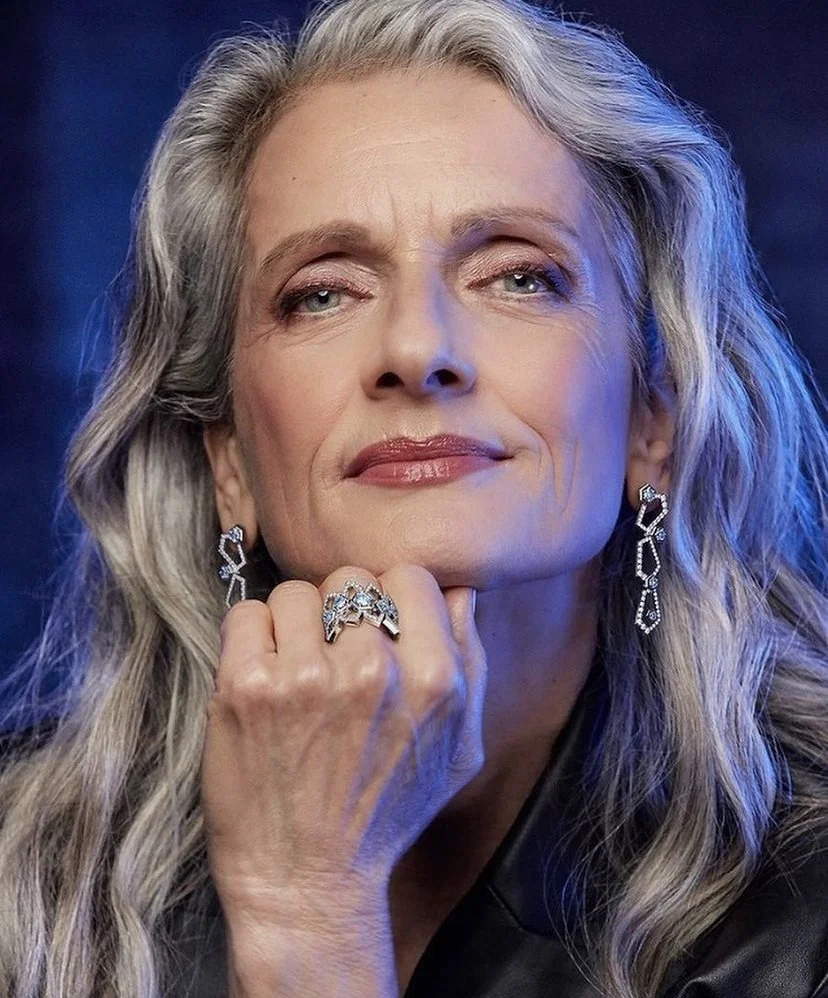 A mature woman with long gray hair, wearing jewelry including earrings and rings, and a black jacket, touches her chin with her hand, looking confidently at the camera. Hair done by Aziza Rasulova for  jewelry photoshoot in NYC.