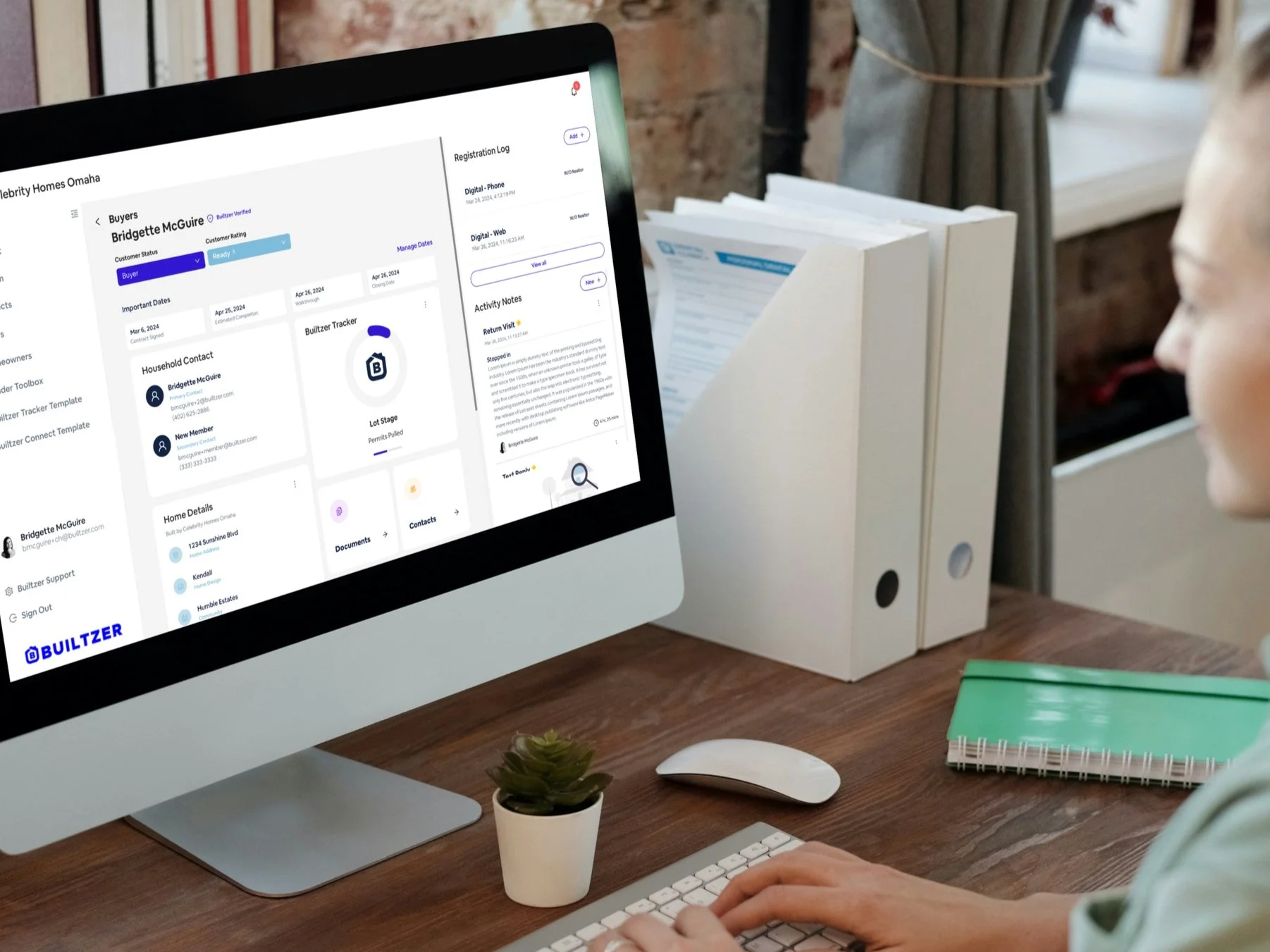 Person working at a desk with an iMac displaying a real estate tracking app, a small potted cactus, a white mouse, a green notebook, and white binders in the background.