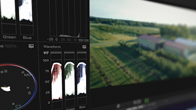 Close-up of a digital audio editing interface on a computer screen, showing waveform and color-coded audio tracks, with a blurred background of a rural landscape visible on another monitor.