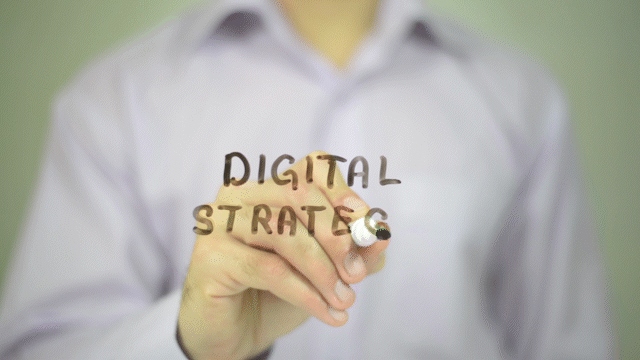 Person in a white shirt writing 'DIGITAL STRATEGY' on a transparent surface with a marker.