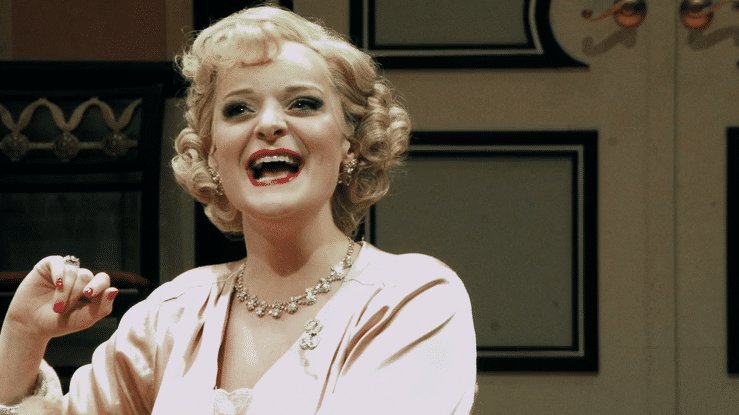 A woman with curly blonde hair smiling and wearing a pearl necklace and a light pink outfit.
