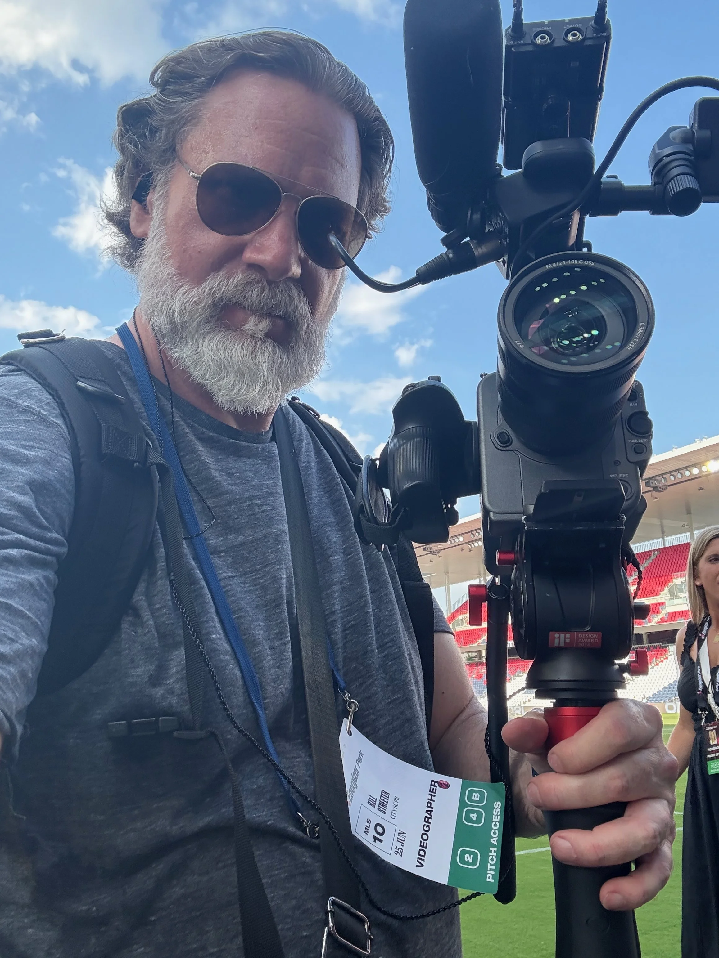 Bill Streeter wearing sunglasses, a gray beard, and brown hair holds a professional video camera at what appears to be a sports stadium or open-air venue under a blue sky with some clouds. He has a VIP or access pass hanging around his neck.