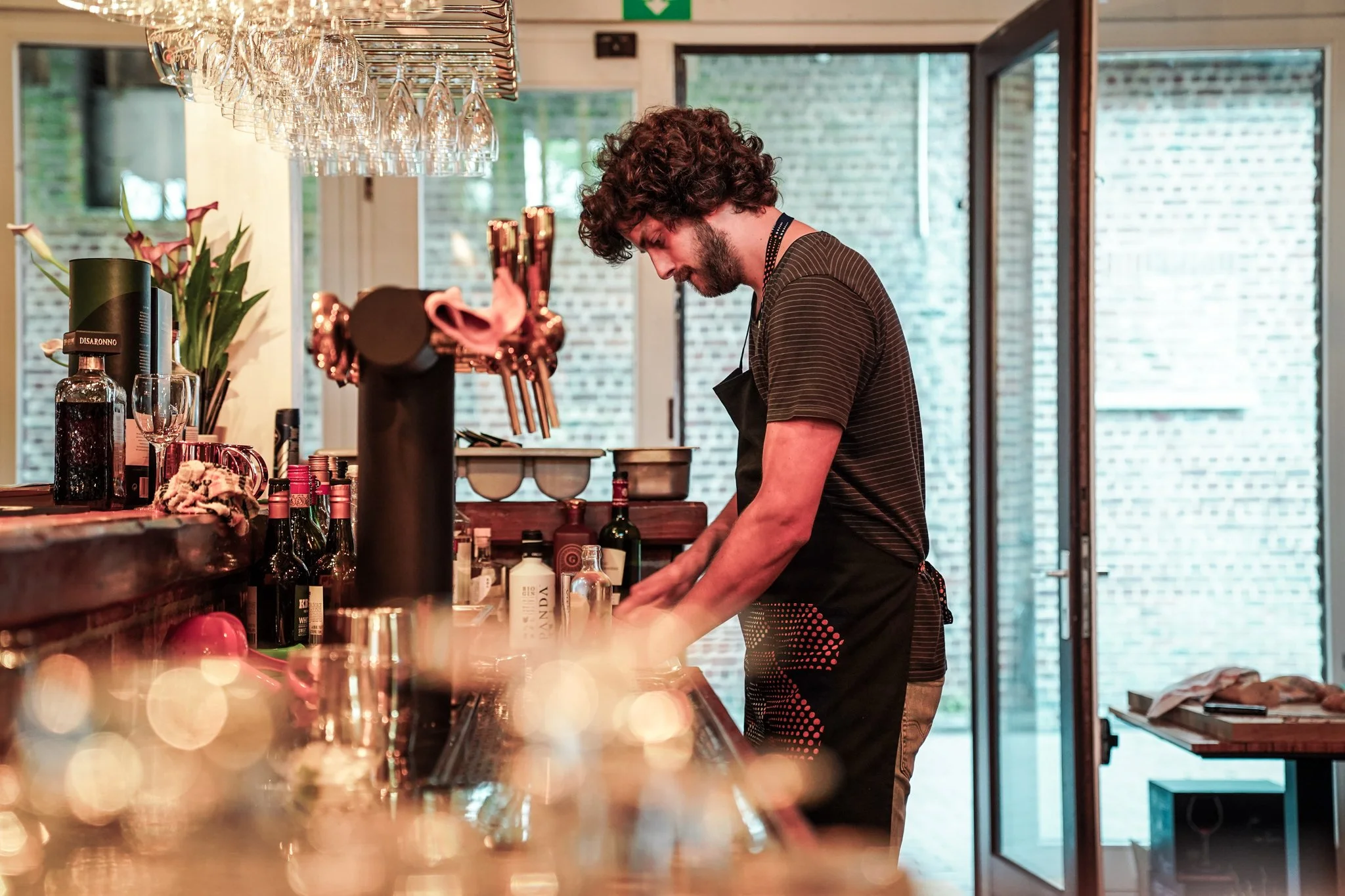 Man bereidt drankjes in een café of bar, omgeven door drankflessen en glazen.