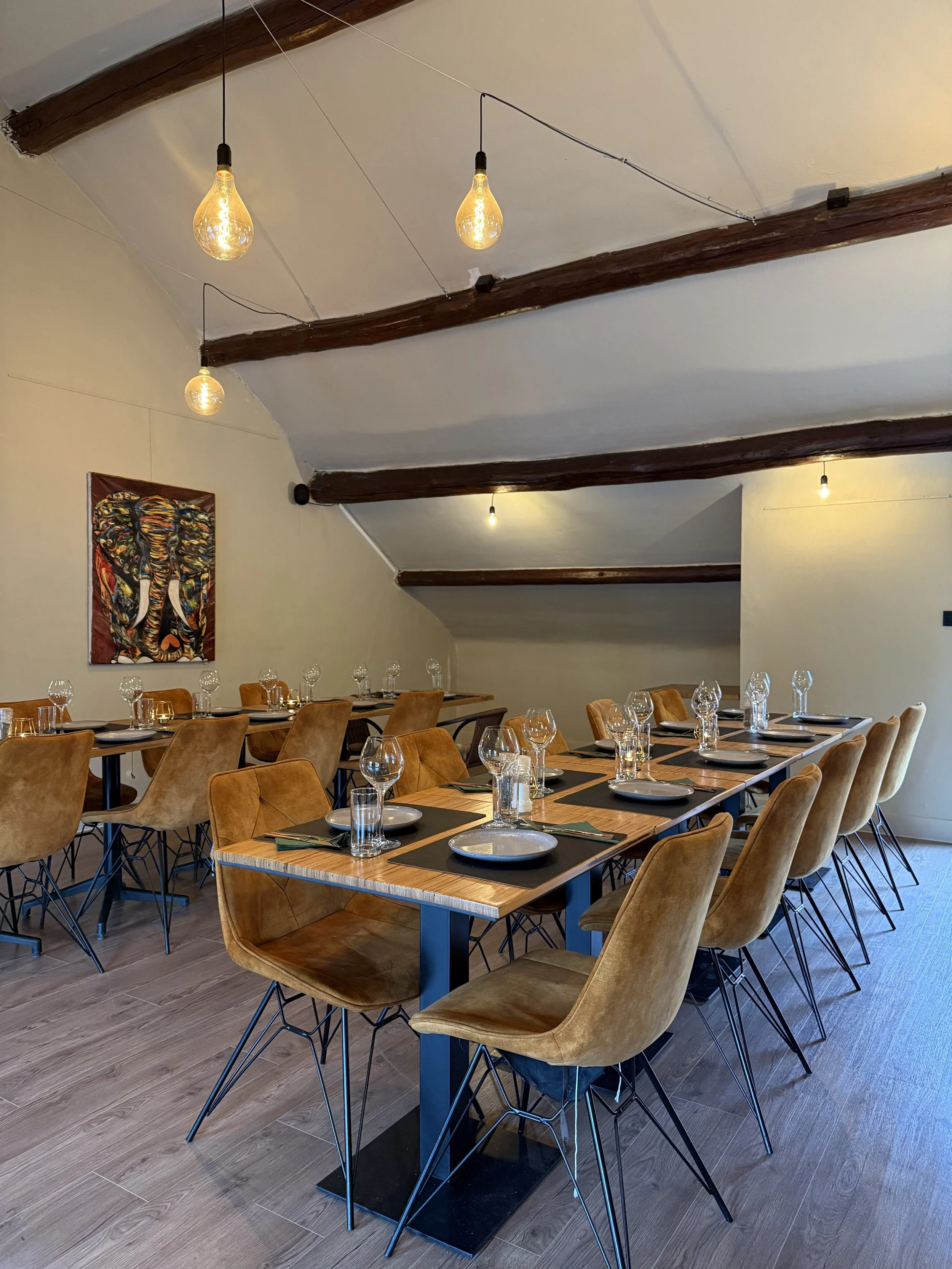 Een restaurantzaal met houten tafels en oranje-bruine stoelen, klaar voor een diner met serveergoed en wijn glazen, verlichte door hanglampen met gloeilampen.