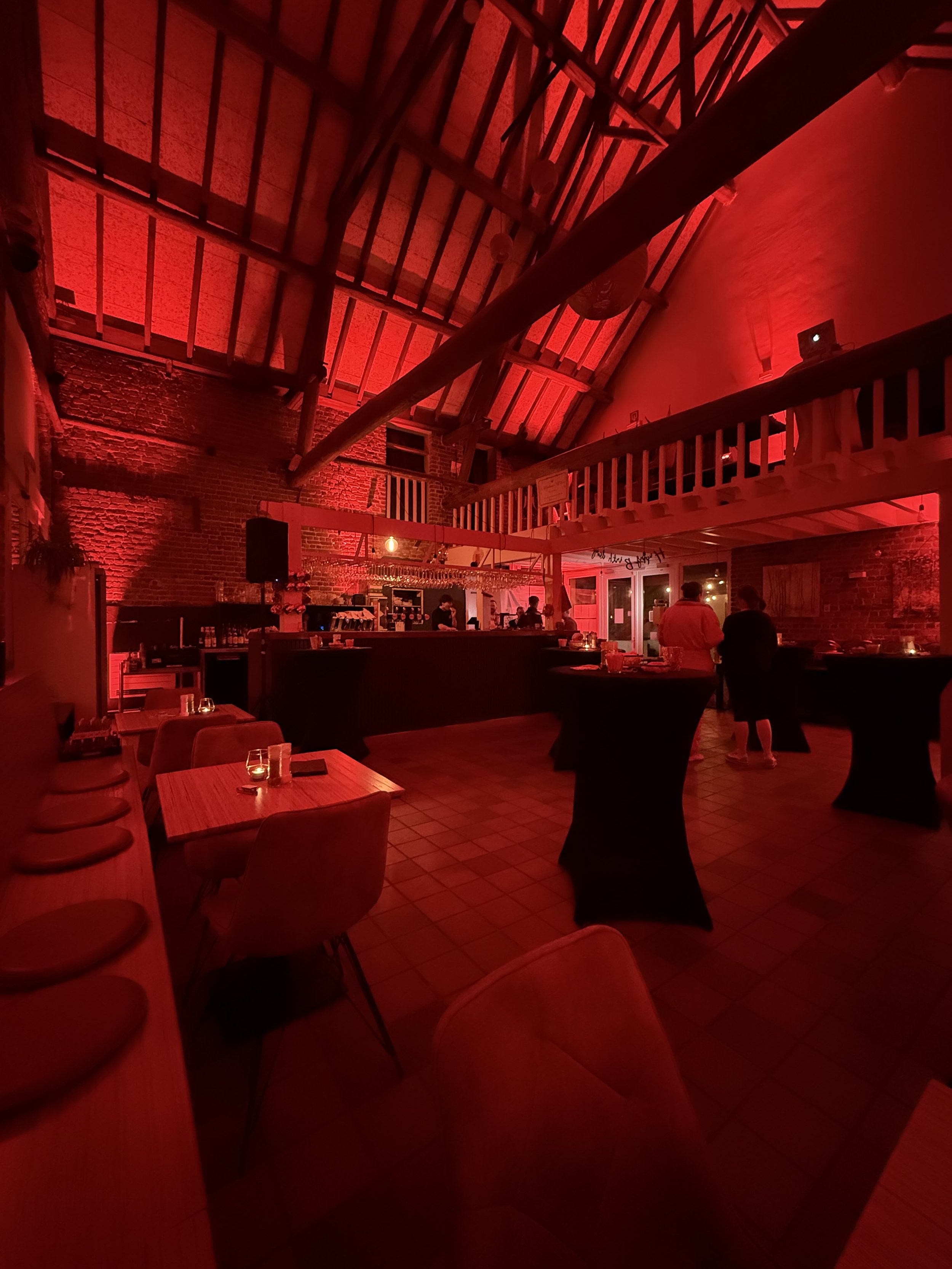 Interieur van het restaurant met rode feestverlichting, tafels met kaarsen, bar met gasten, en een hoog plafond met houten balken en een balkon.