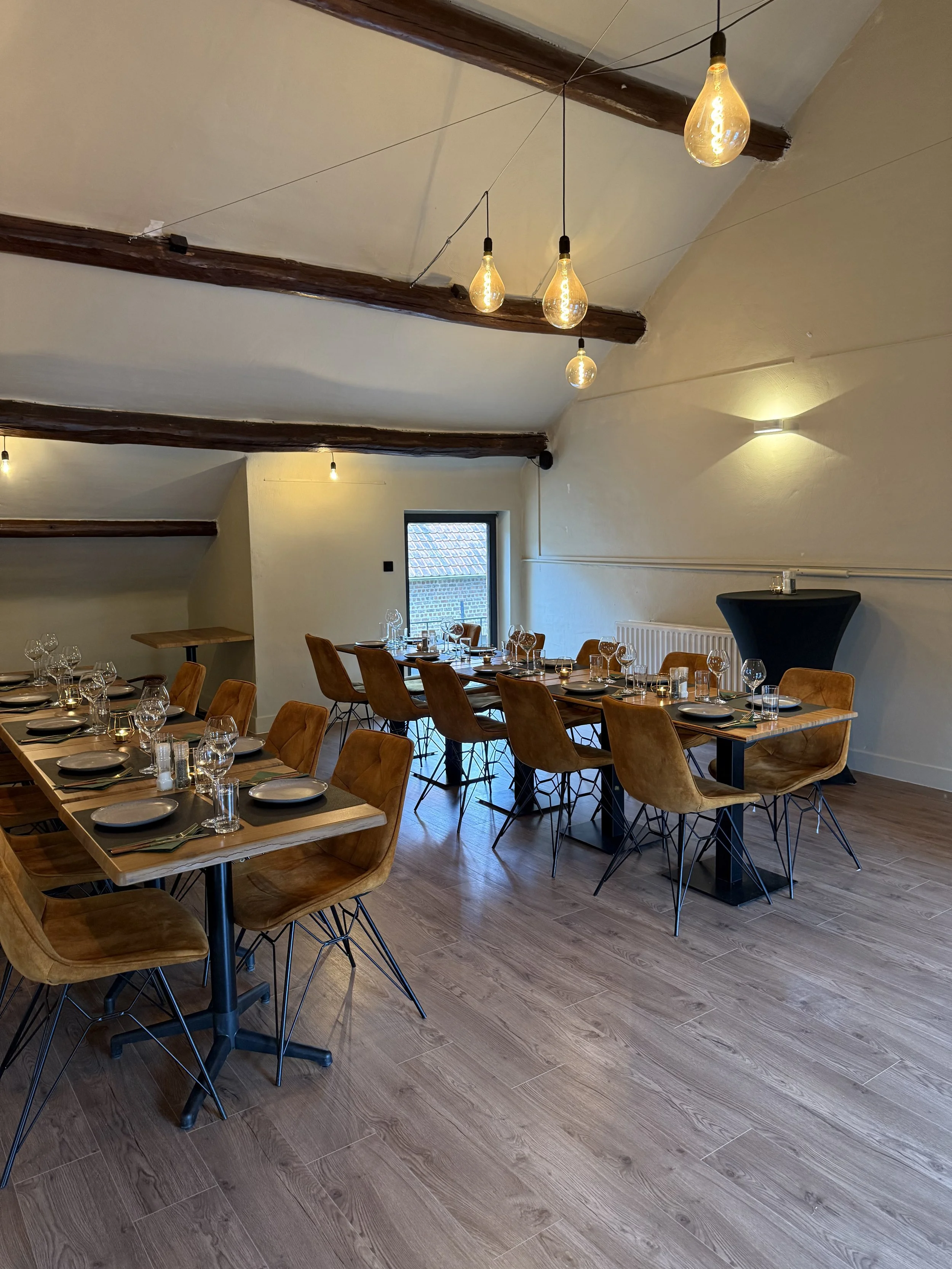 Een gezellige restaurantruimte met houten tafels, oranje bruin beklede stoelen, wijn- en waterglazen, borden en bestek, een grote venster en decoratieve lampen aan het plafond.