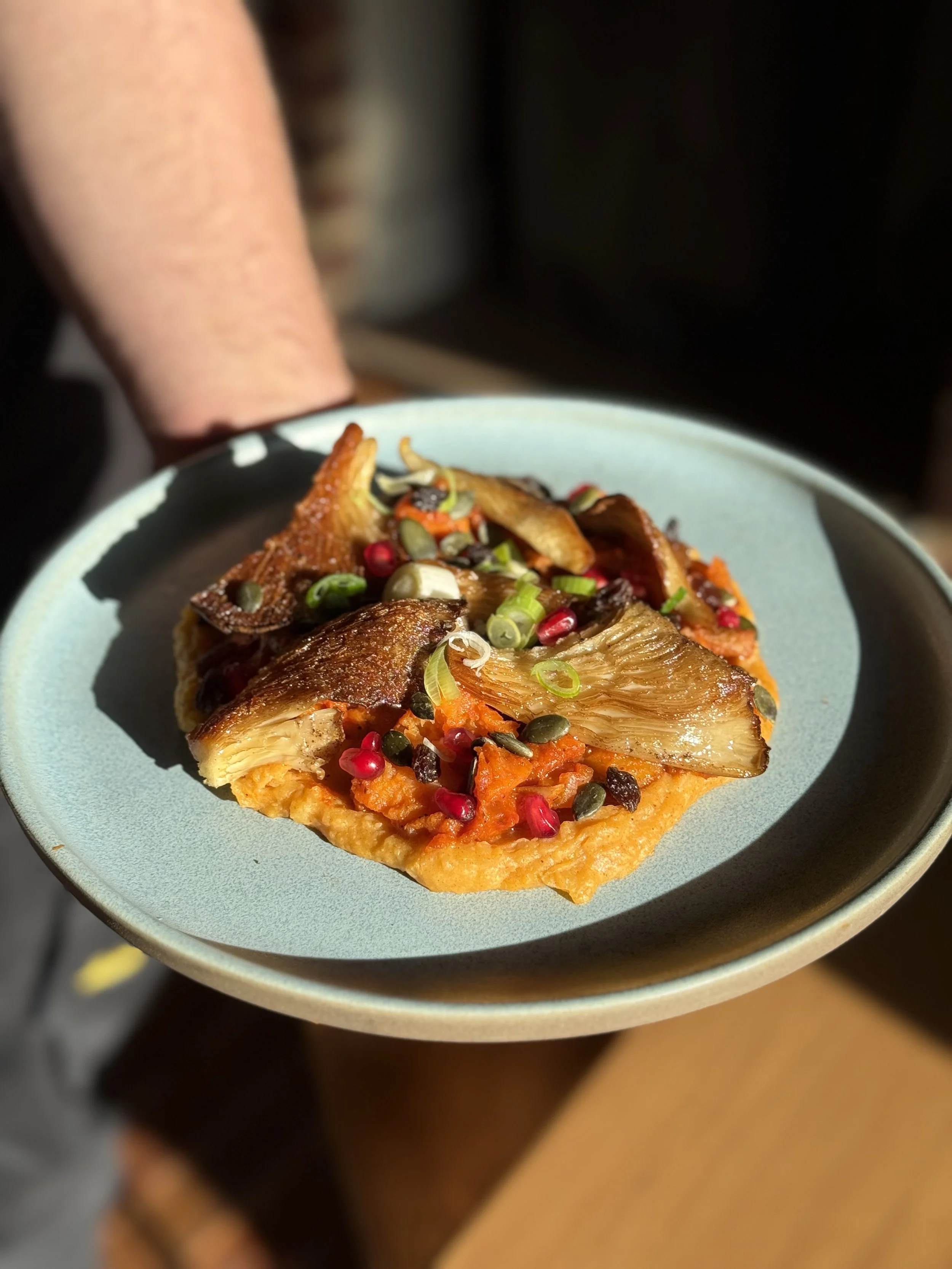 Gerecht met gebakken oesterzwammen op een bedje van puree met een getomateerde saus, geserveerd op een bord met zonlicht erop, versierd met fijn gesneden lente ui en granaatappelpitten.