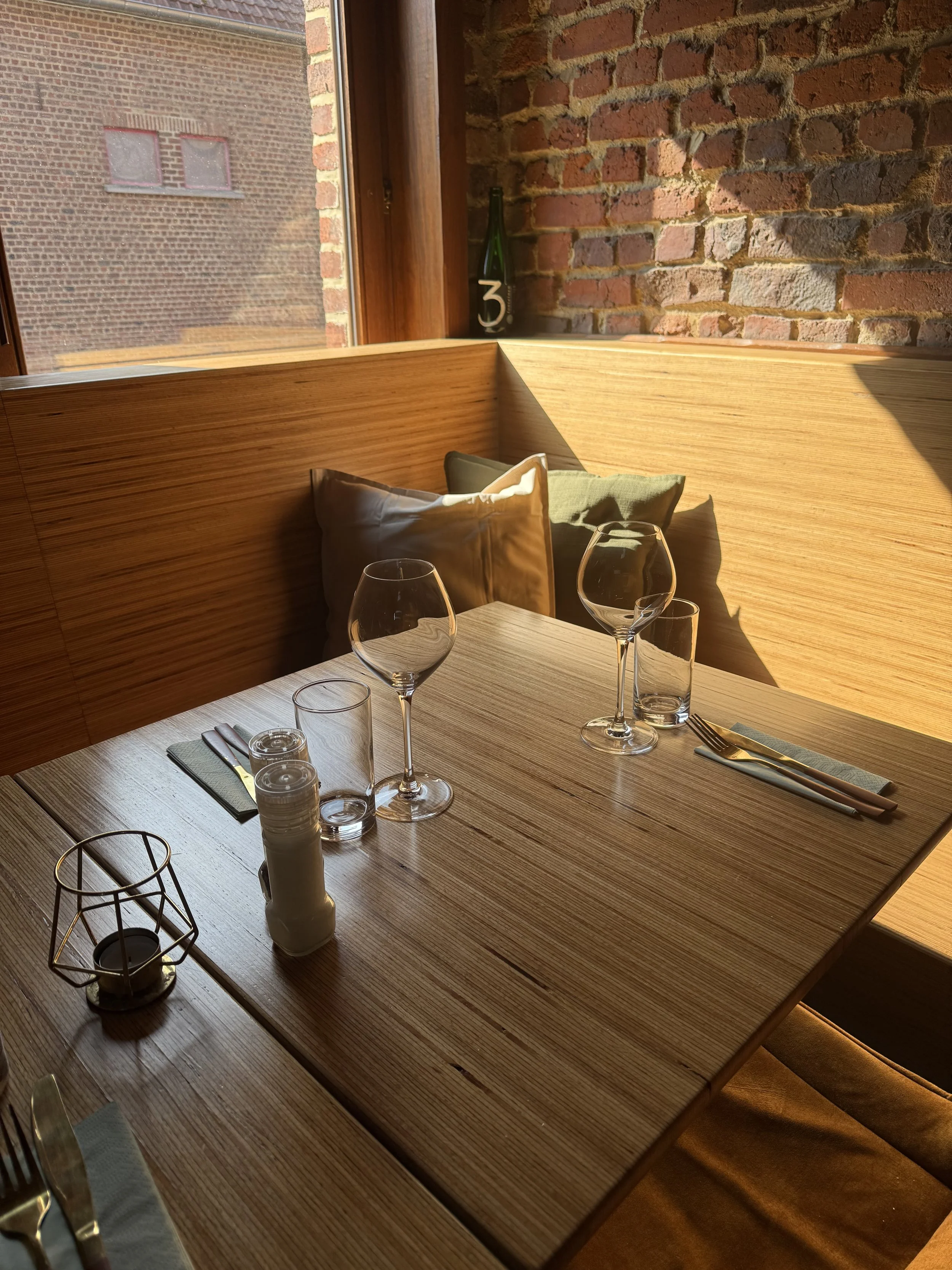 Een gedekte eettafel in het restaurant met twee lege wijnglazen, bestek, een klein kaarsje en zout- en peperstel. De tafel staat bij een raam met veel zonlicht en een bakstenen muur op de achtergrond.