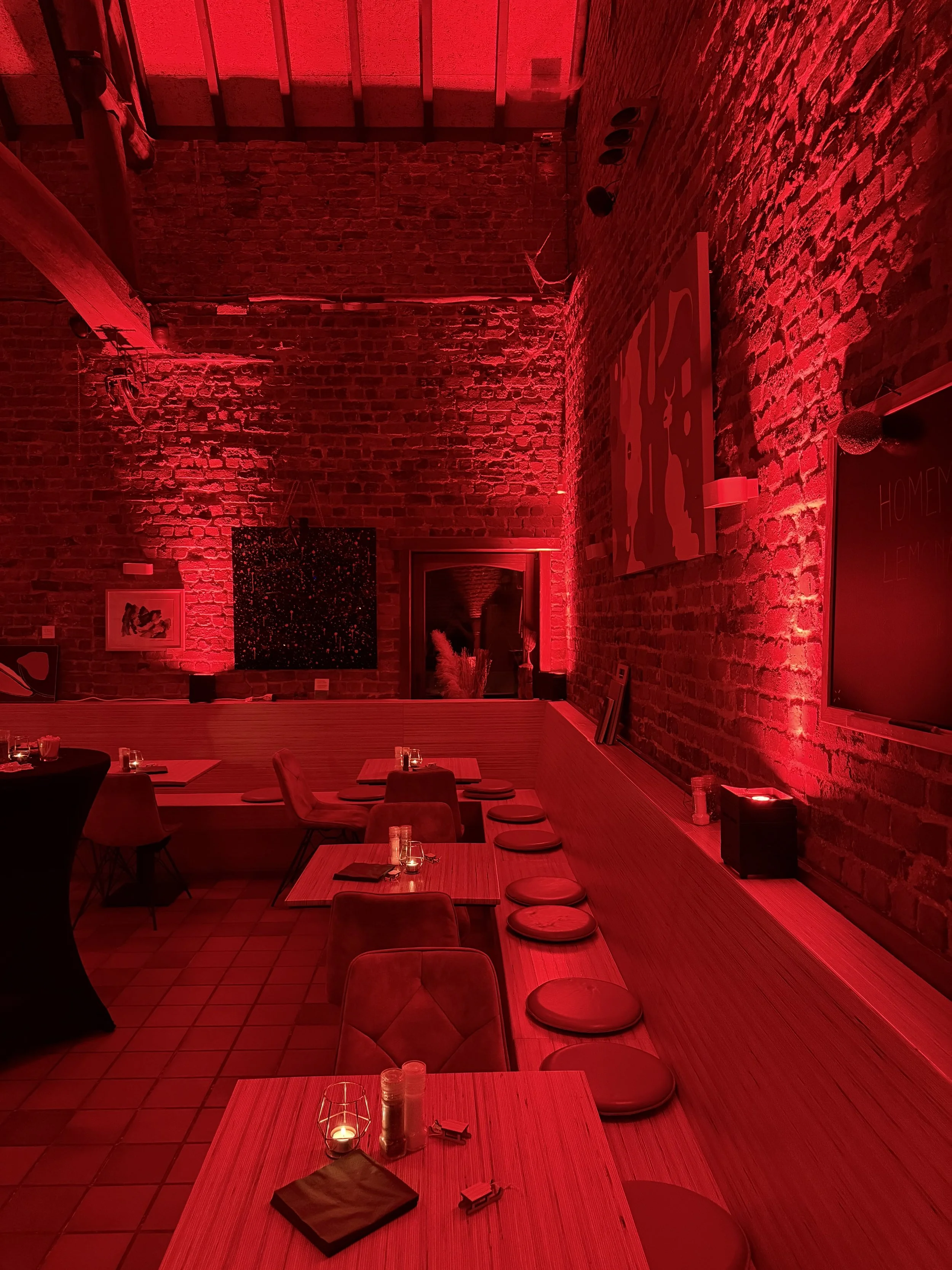 Binnenkant van een restaurant met rode verlichting, houten tafels met placemats, kaarsen en bestek, en rode stoelen, met blootgelegde bakstenen muren.