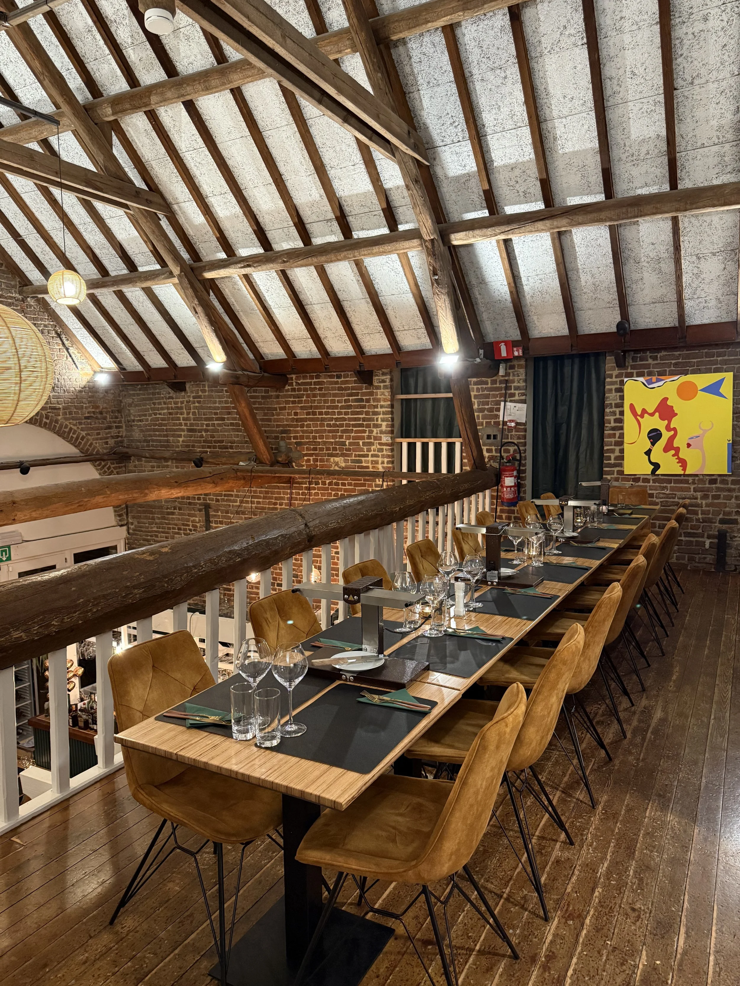 Eetafel in een restaurant met houten tafel, gele stoelen, glazen en bestek, in een ruimte met een houten plafond en bakstenen muren, en moderne kunst aan de muur.