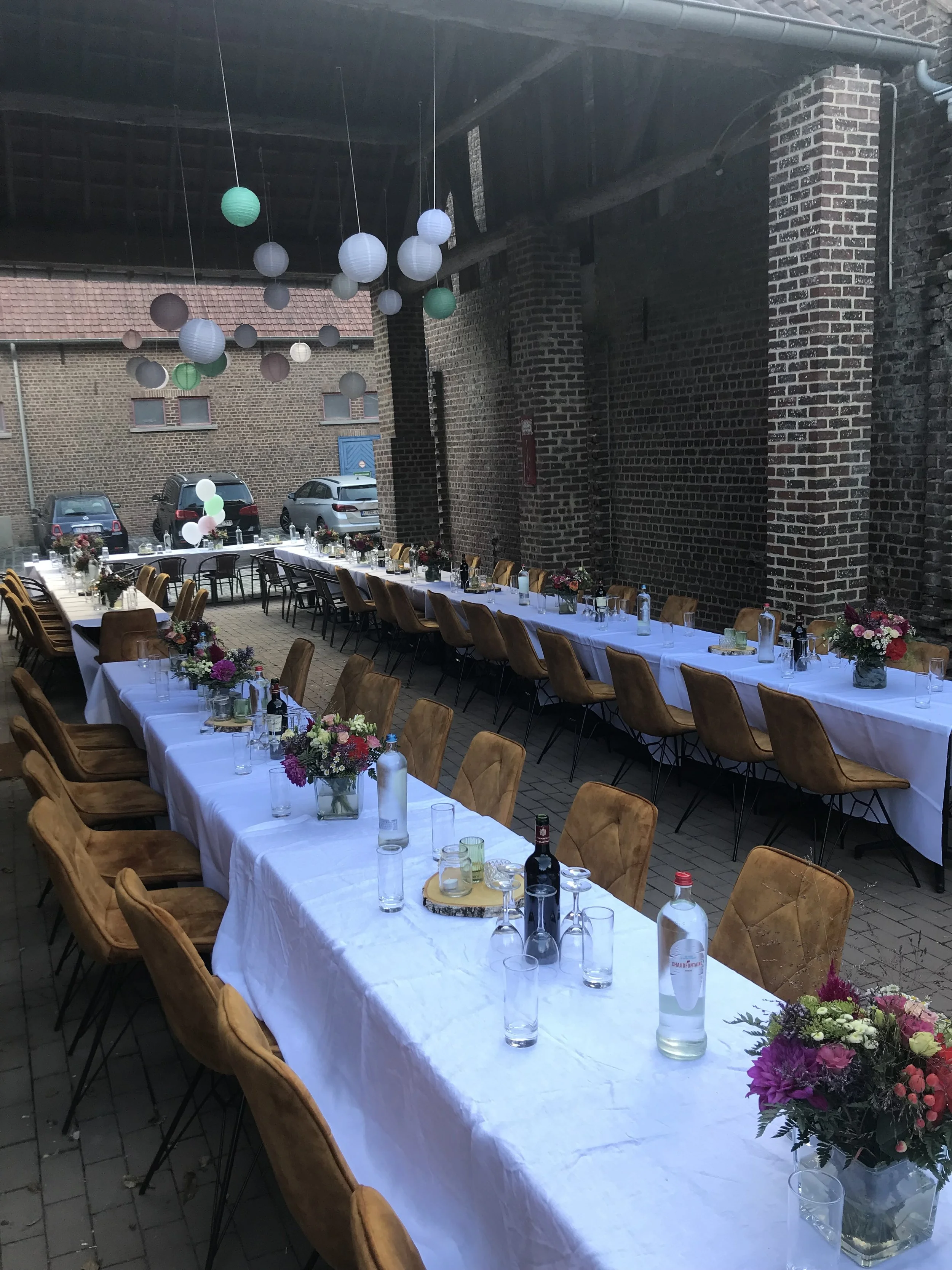 Een feestzaal ingericht voor een evenement met lange tafels bedekt met witte tafellakens, versierd met bloemstukken en drankjes, met papieren lantaarns en ballonnen in de bovenruimte.