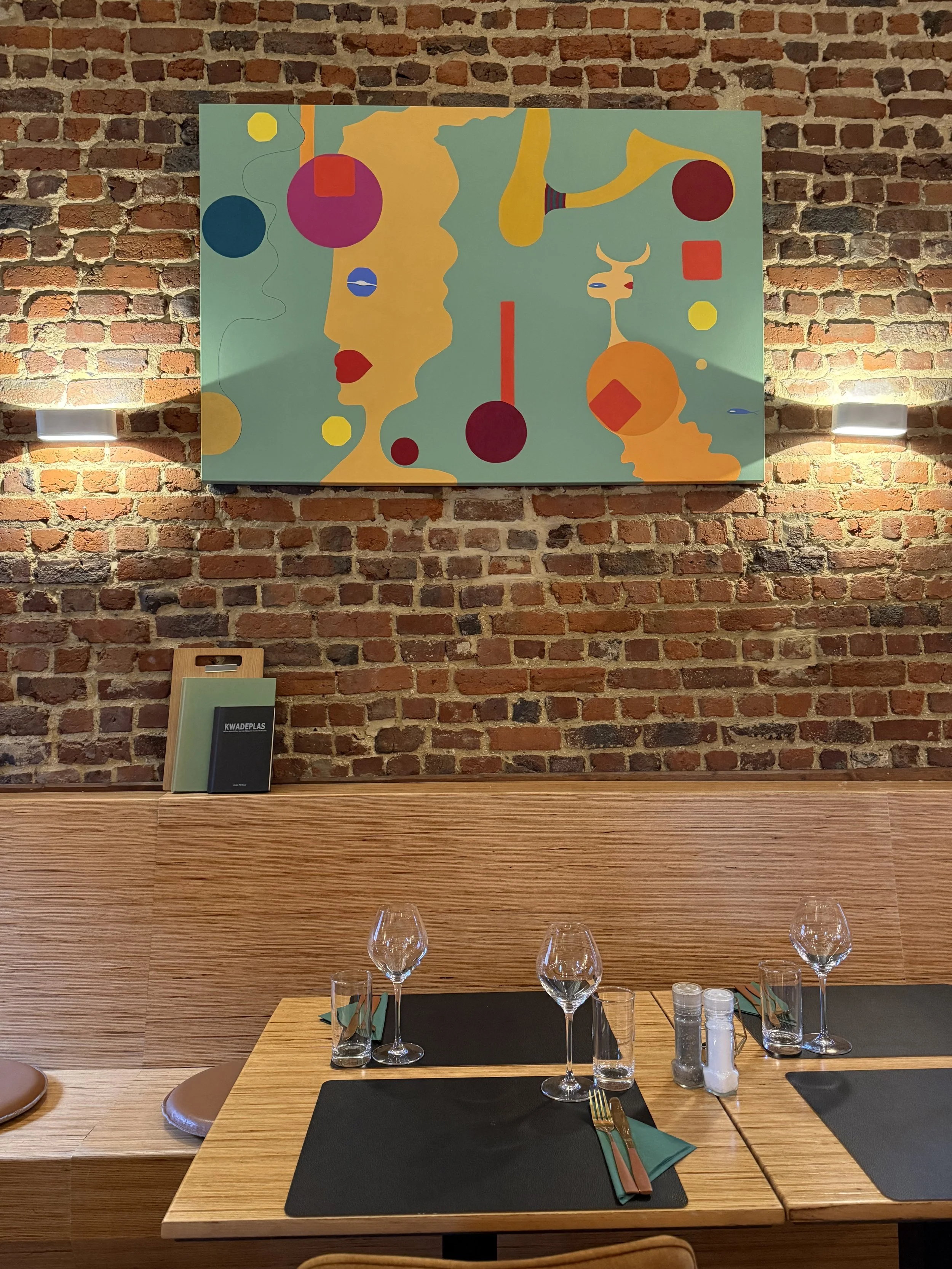 Een restauranttafeltje met vier lege wijnglazen, bestek, en zwart placemats tegen een houten bank, met een bakje zout, peper en suiker op de tafel. Op de bakwand hangt een abstract kunstwerk met geometrische en menselijke silhouetten tegen een bakste