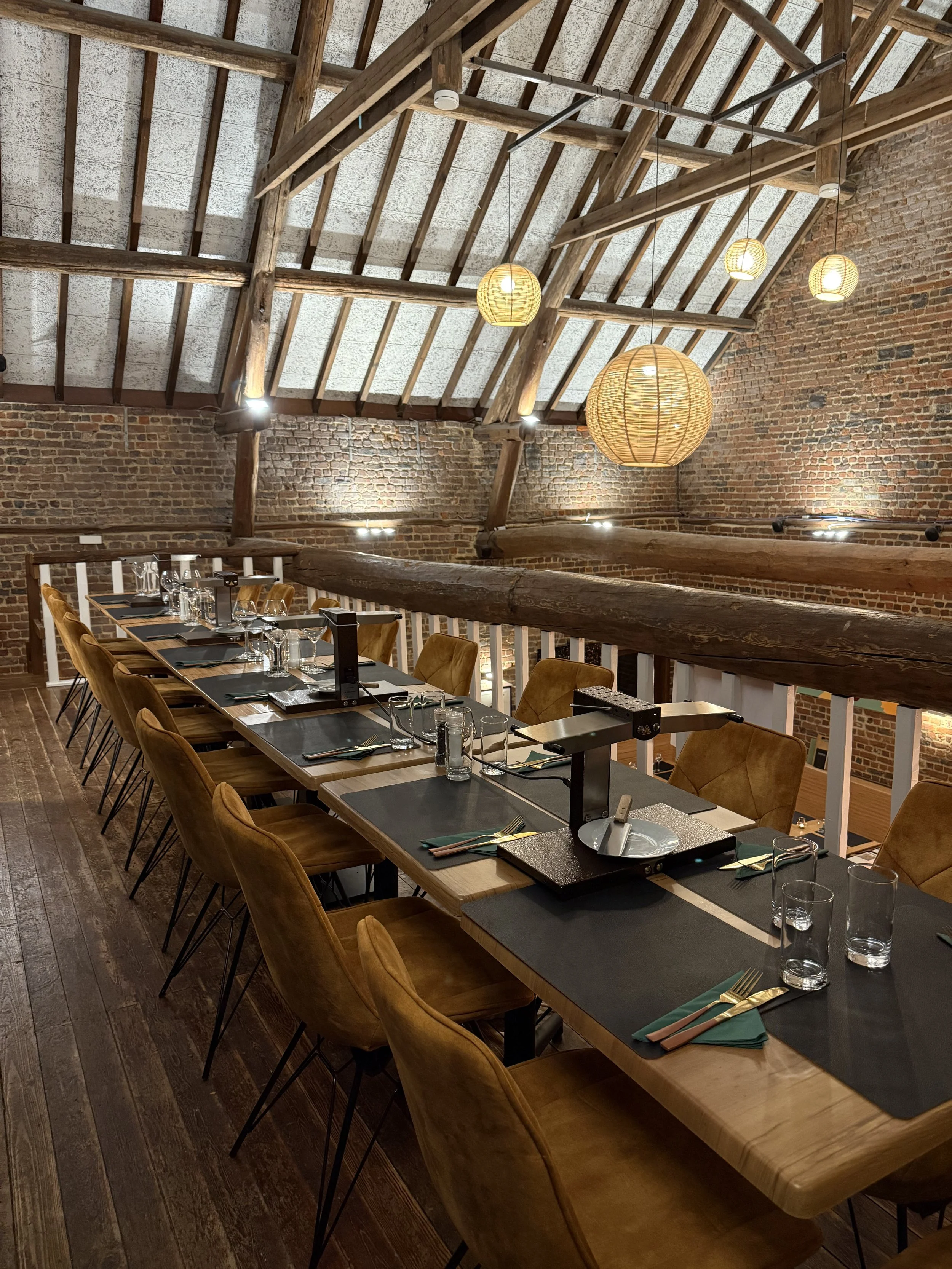 Een restaurant of eetzaal met een houten tafel op de bovenverdieping, sierlijke bruin-beige stoelen, open bakstenen muren, houten balken en hanglampen in een rustieke stijl.