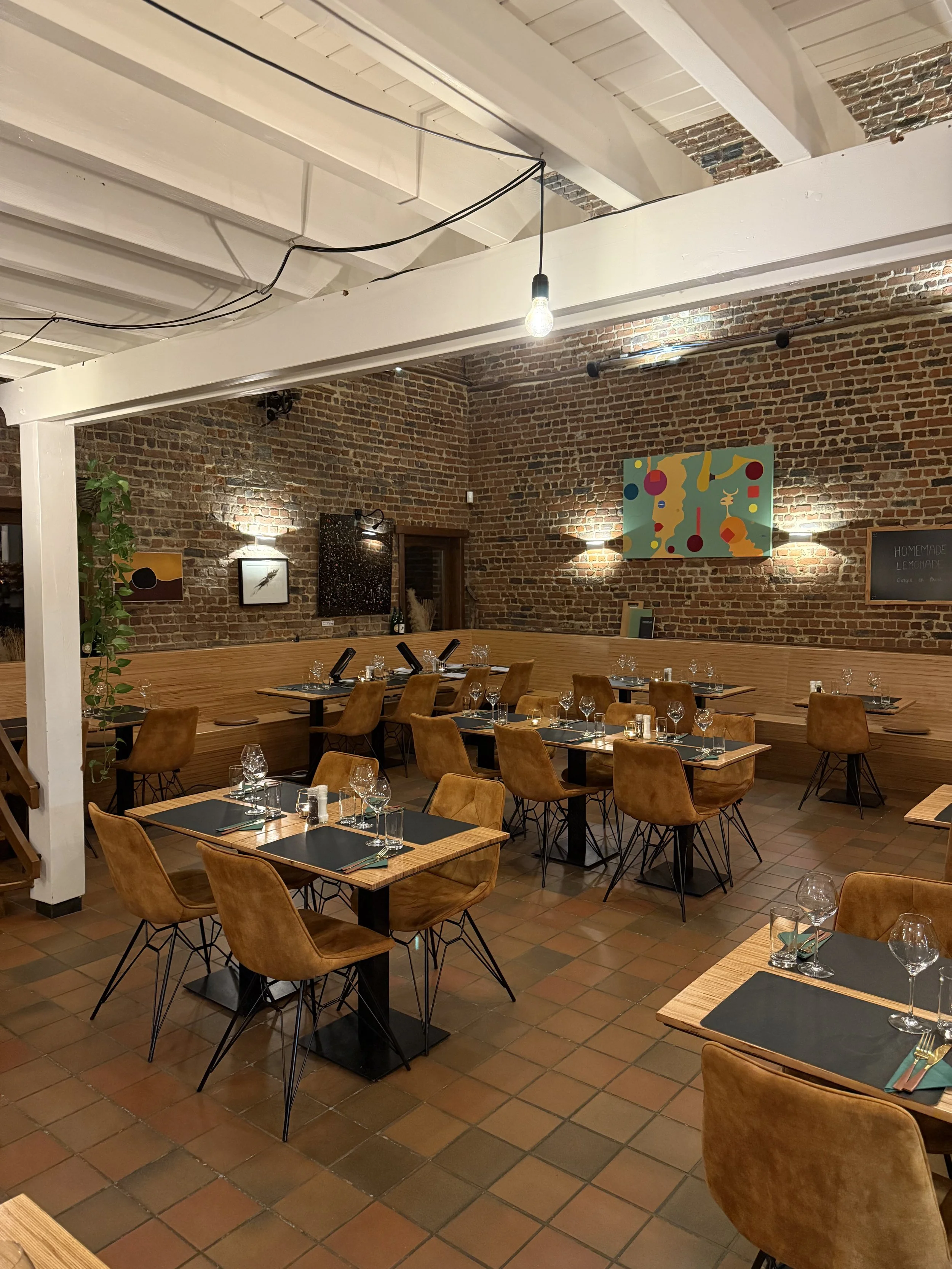 Een uitnodigende restaurantzaal met houten tafels en leren stoelen, gedekte tafels met wijnglazen en bestek, muurversiering met kunstwerken, brick muren en een industrieel plafond.