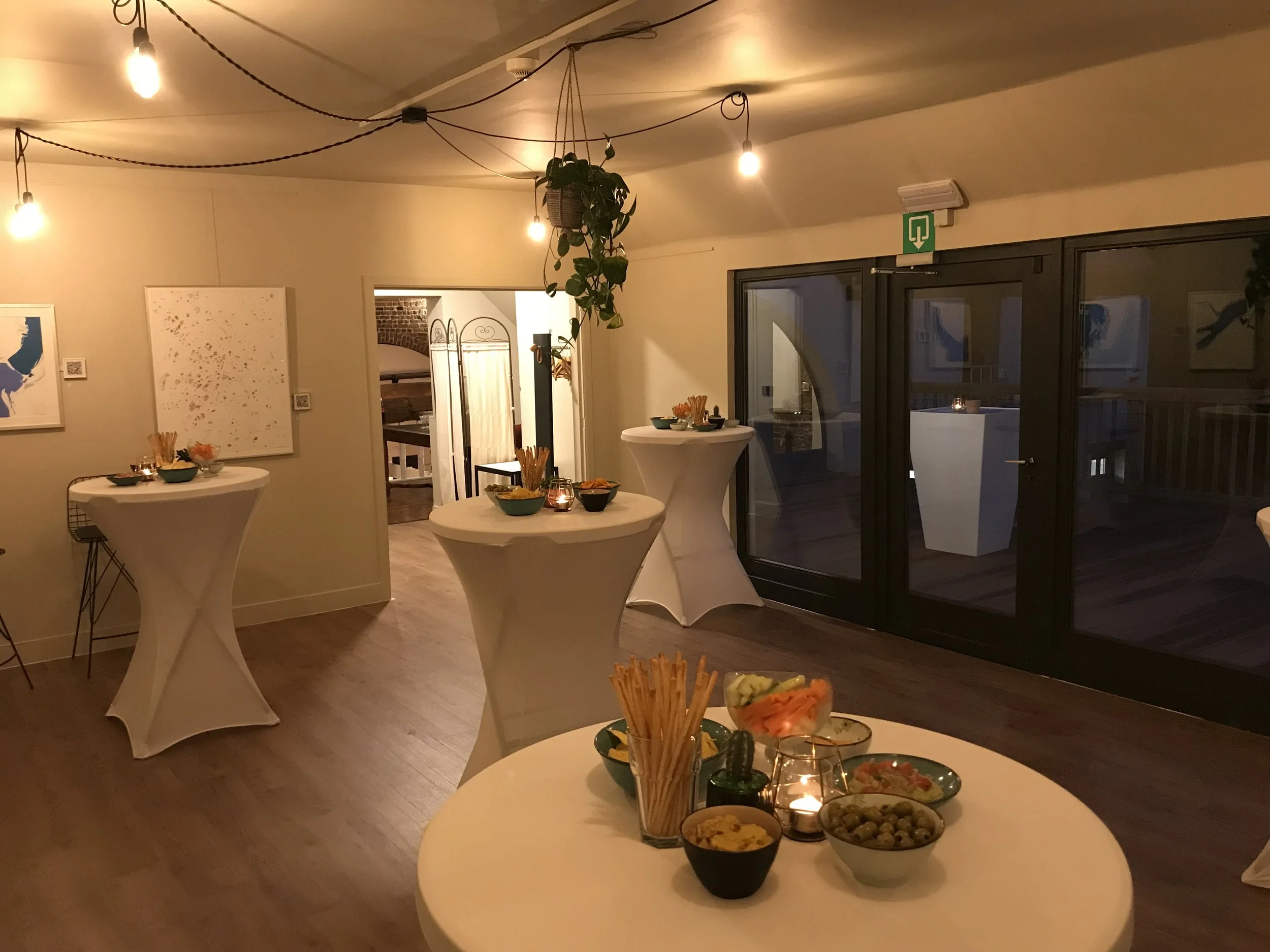 Interieur van een feestruimte met hoge cocktailtafels bedekt met witte tafelkleden, snacks en kaarsen, decoratieve verlichting, en een plantenrek hangend aan het plafond.