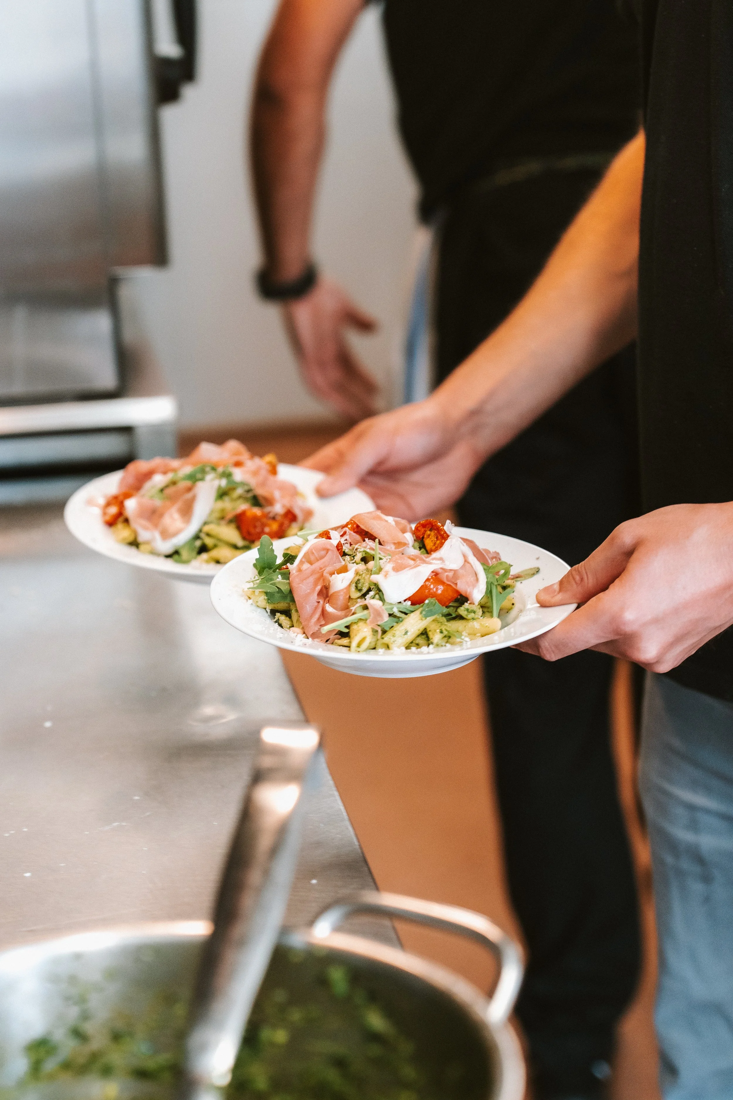 Persoon die twee borden pasta met ham, kerstomaten, en rucola vasthoudt in de keuken.