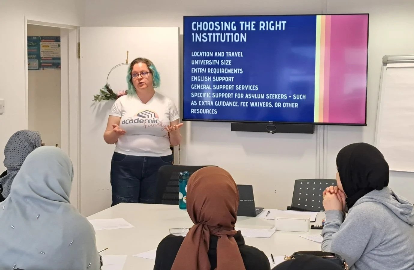 A group of women sit in a white room, watching the presenter (Dr. Carly McNamara) deliver a workshop on Further and Higher Education. The Text on the screen reads: Choosing the Right Institution