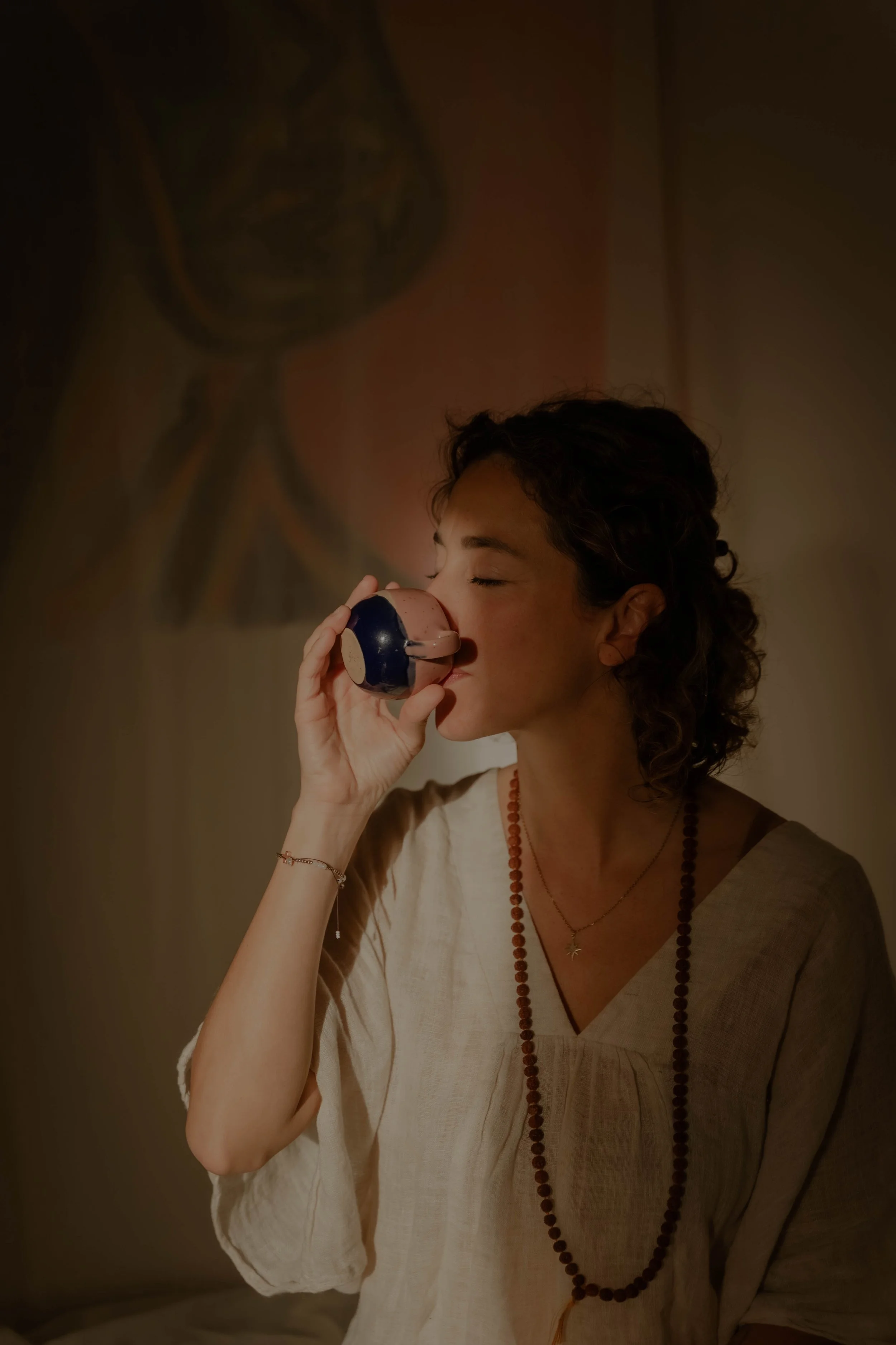 A woman with curly hair, wearing a beige top, a beaded necklace, and bracelets, is holding a cup to her lips with her eyes closed, standing in front of a warm-colored wall with a painted design.