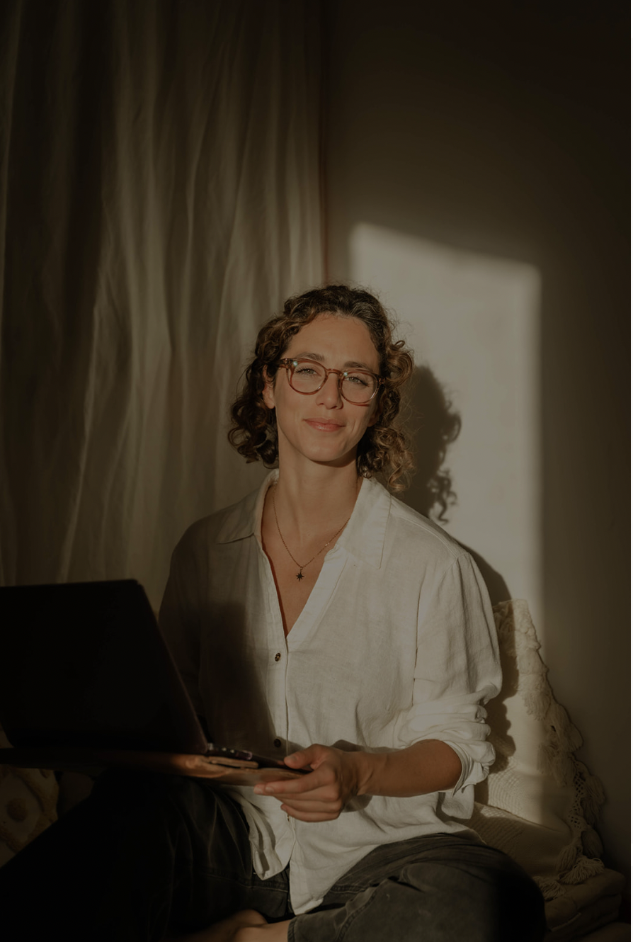 A woman with curly hair and glasses sitting on a couch with a laptop, sunlight casting a shadow on the wall behind her.