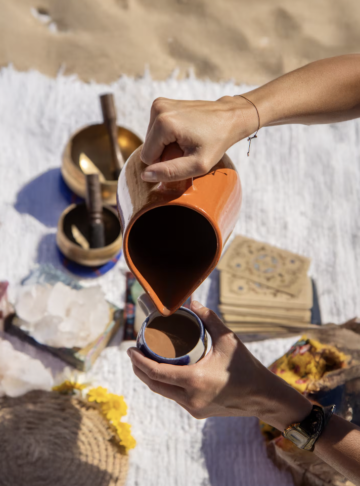 Two people are pouring coffee from an orange teapot into a mug during a beachside gathering. The scene includes a white blanket, wooden bowls with spoons, a stack of paper napkins, and some flowers.