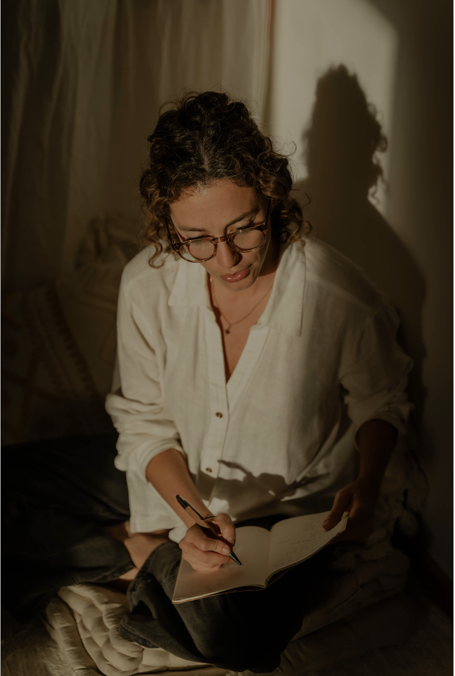 A woman with curly hair and glasses is sitting on a bed, writing in a notebook with a black pen. She is wearing a white shirt and appears to be in a dimly lit room.