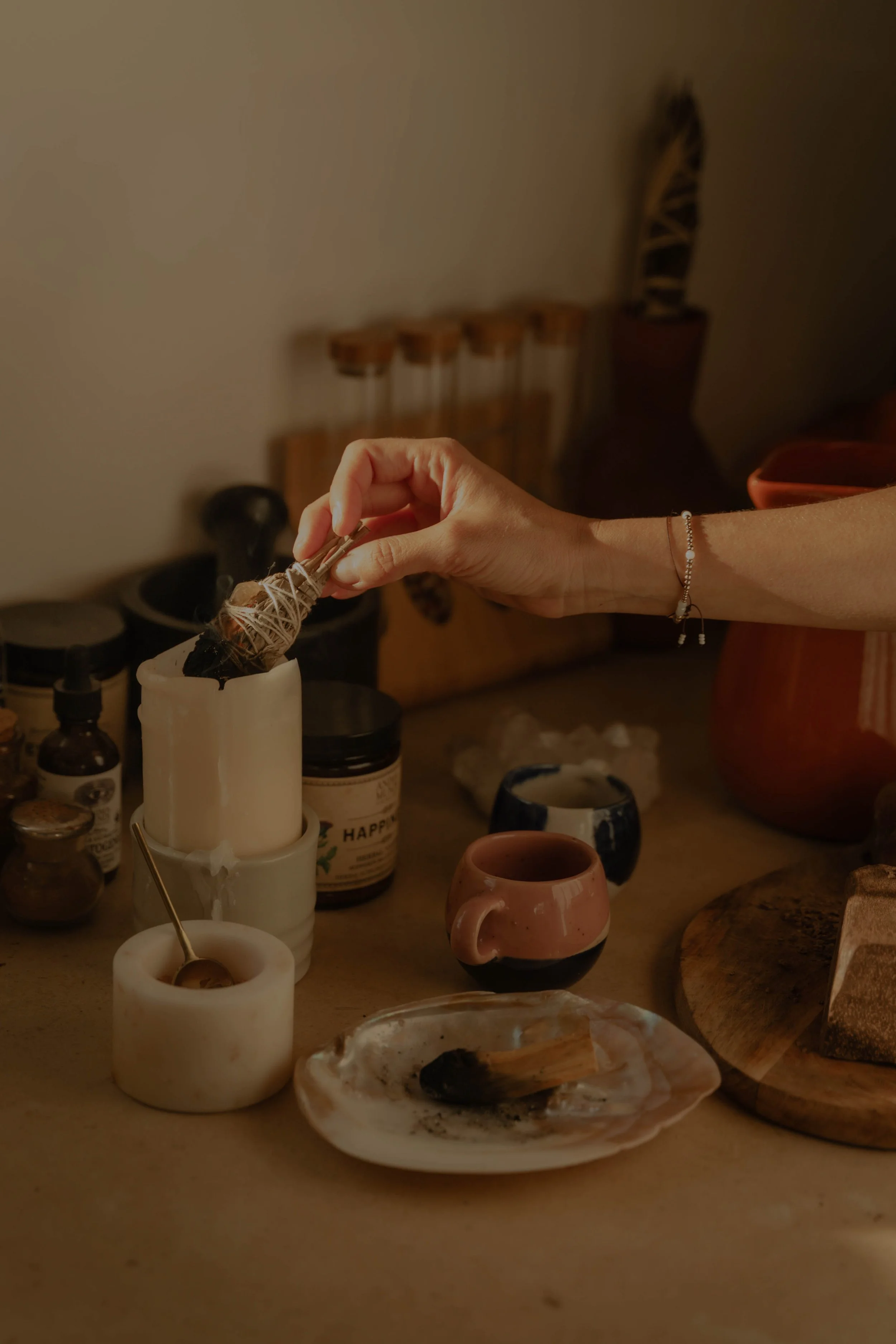 A person is abalone a crystal with thread and a needle while surrounded by various candles, jars, and pottery on a wooden table in a cozy, dimly lit space.