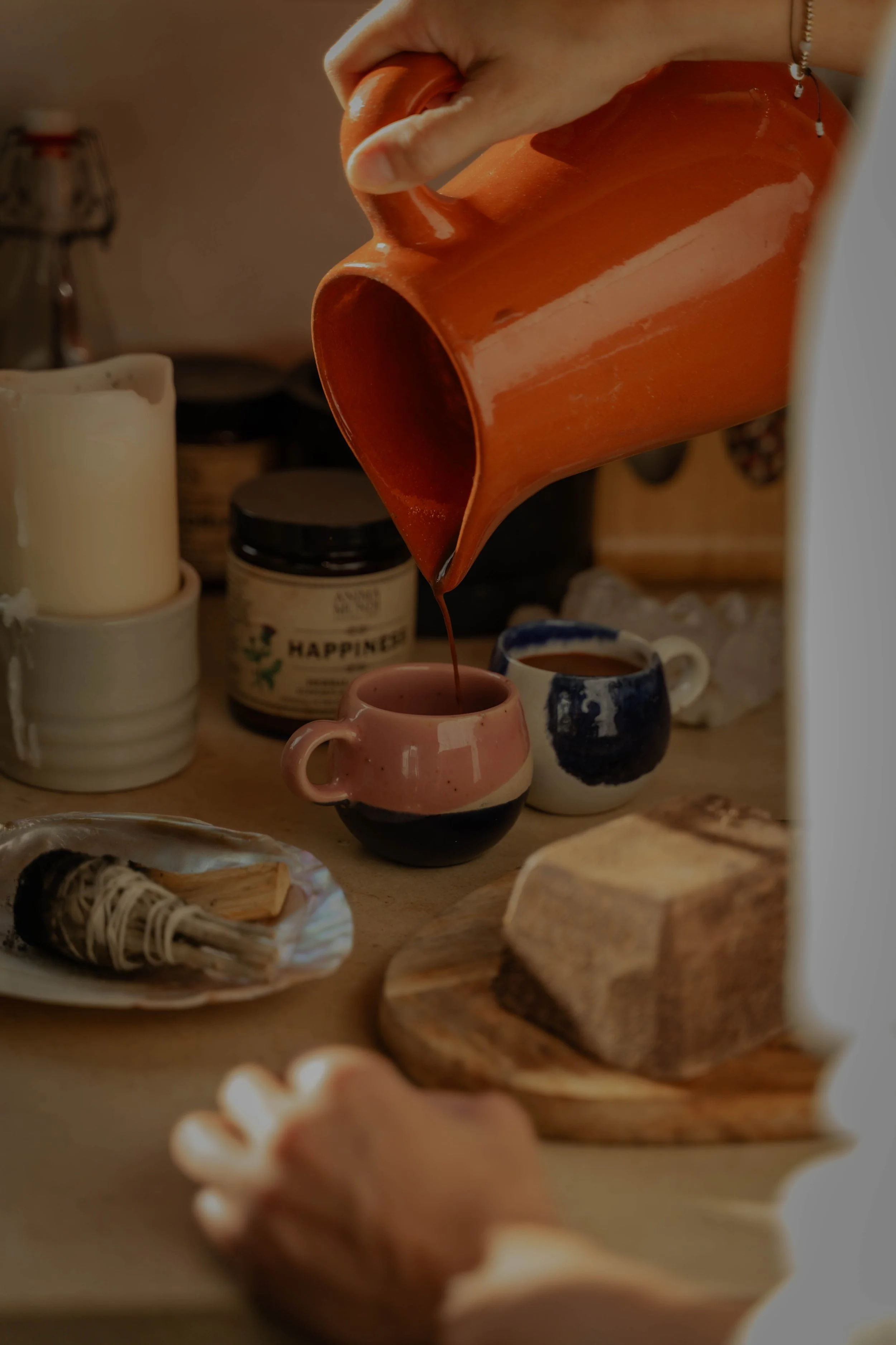 Person pouring liquid from a large orange mug into a small pink and black mug on a wooden table.