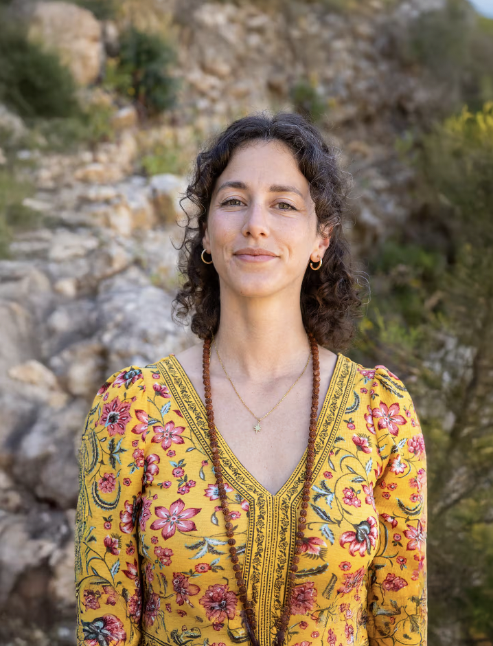 A woman with dark curly hair, light skin, dressed in a yellow floral dress, outdoors surrounded by rocks and greenery.