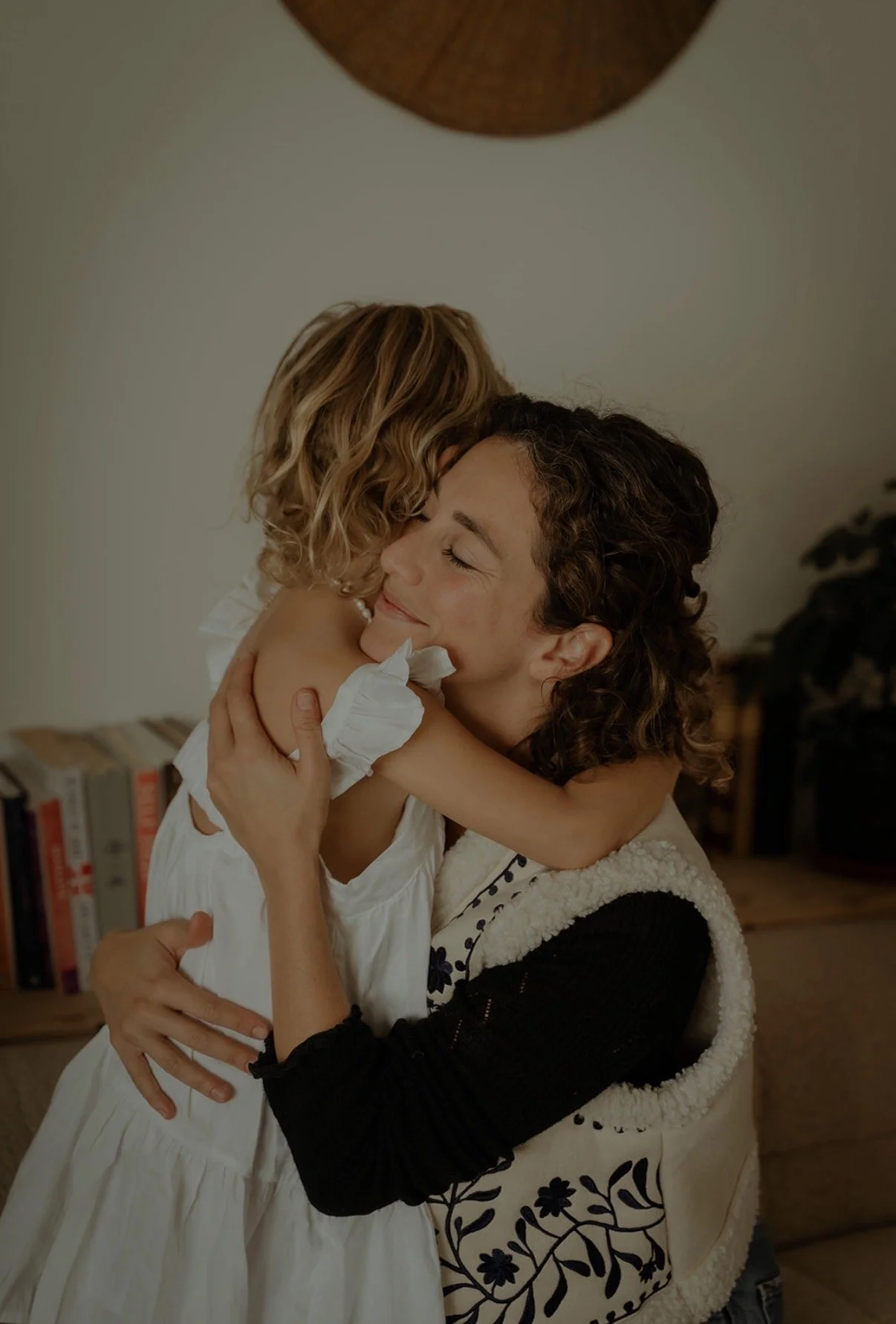 Two women, one with curly brown hair and the other with blonde hair, are embracing each other with joy and affection in a cozy indoor setting.