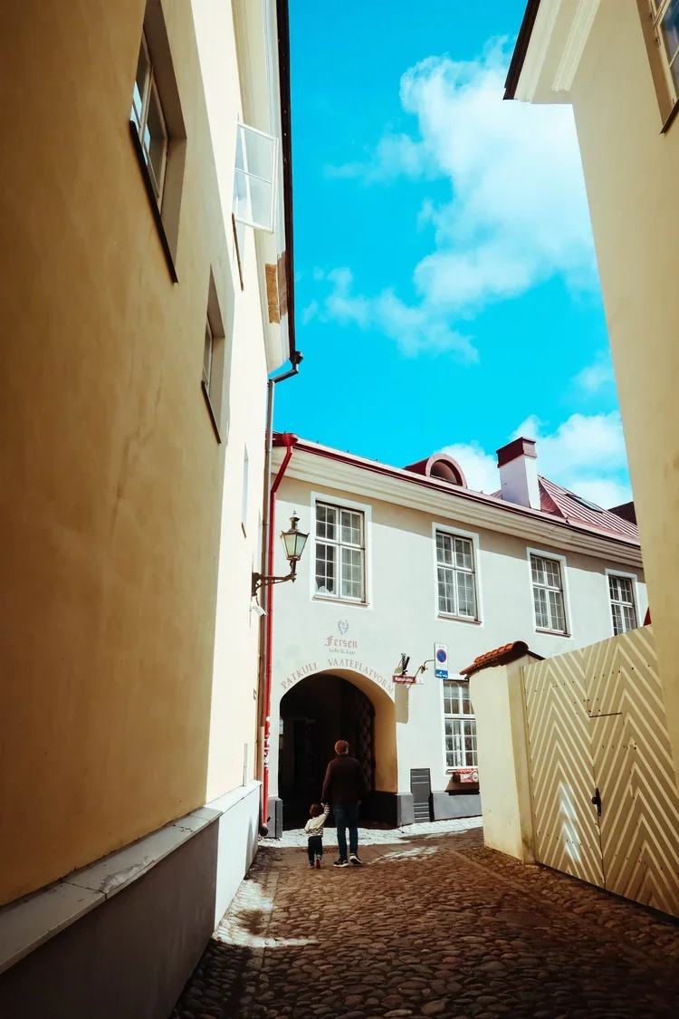 A narrow cobblestone alleyway between yellow and white buildings, with a person and a child walking away.