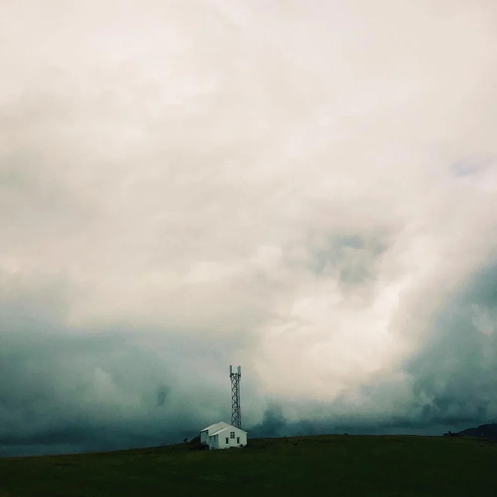 A small white house with a tall communication tower nearby on a grassy hill under a cloudy sky.