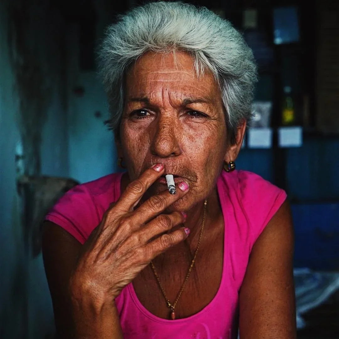 A woman with short gray hair wearing a pink sleeveless shirt, holding a cigarette near her mouth, and looking at the camera.