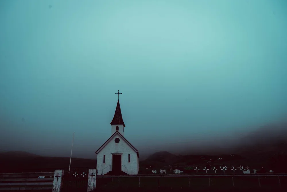 A small white church with a steeple and cross on top, situated in a dark, misty landscape with hills in the distance.