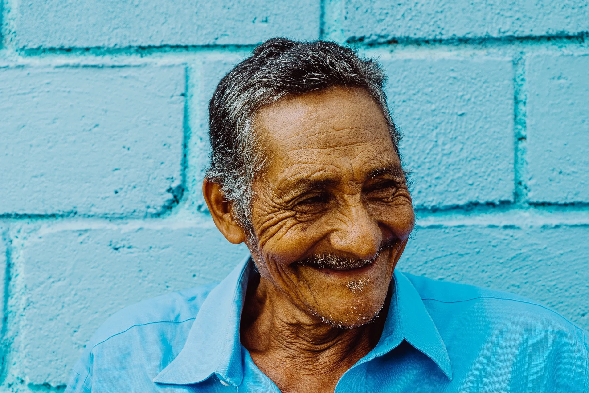 Close-up of an elderly man with gray hair smiling, wearing a light blue collared shirt, standing against a bright blue cinder block wall.