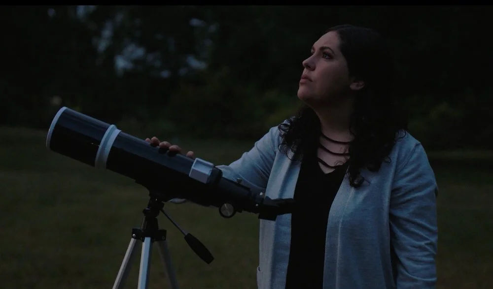 A woman standing outside at dusk looking through a telescope.