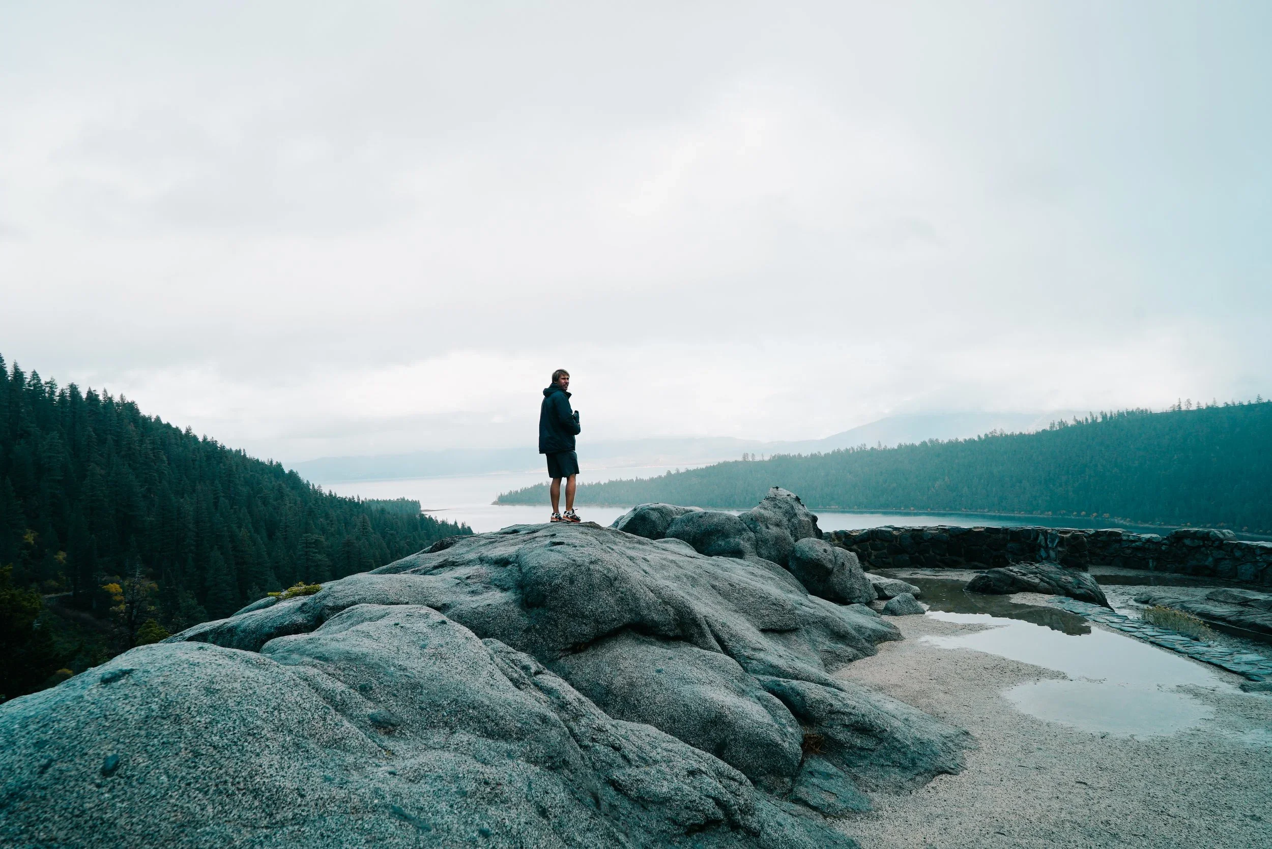 A person standing on rocks by a body of water, surrounded by forested hills and an overcast sky.