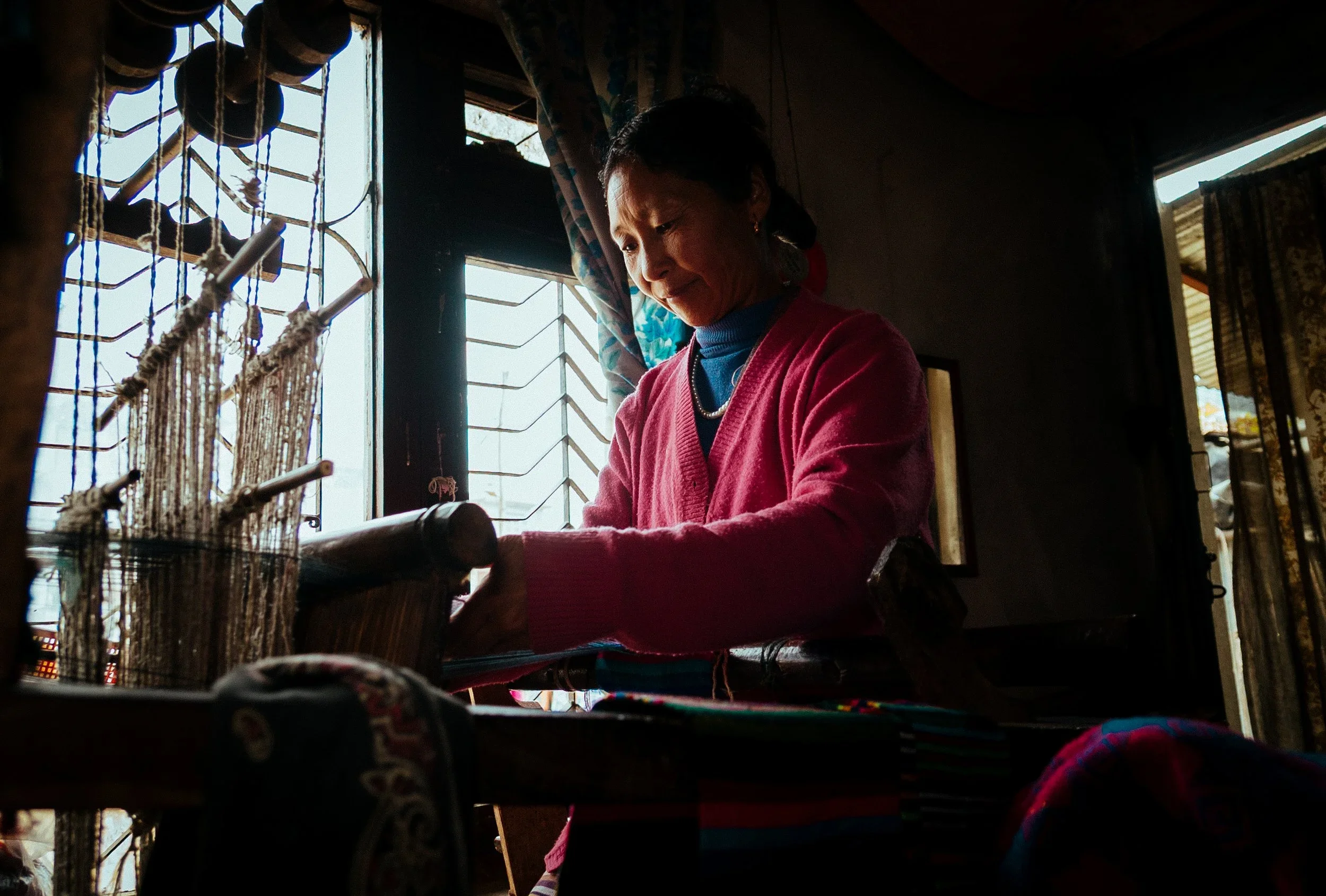 A woman in a pink sweater working on a loom inside a room with open window shutters.