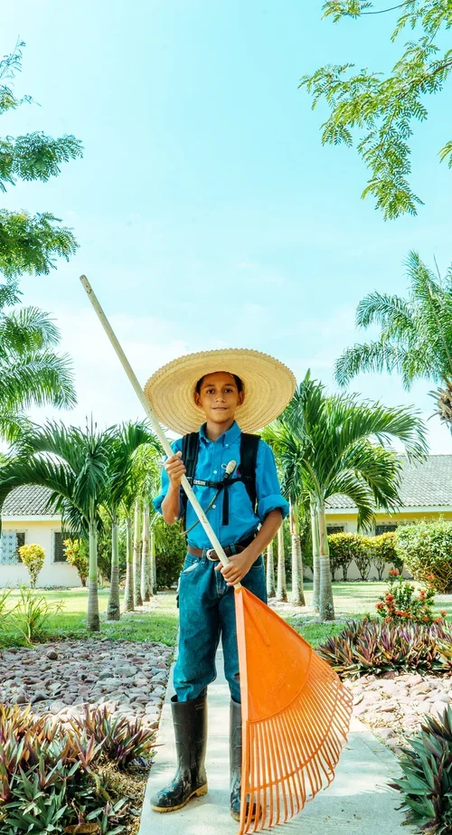 Young boy with a backpack, wearing a large straw hat, blue shirt, jeans, and black boots, holding a leaf rake outside in a garden with palm trees.