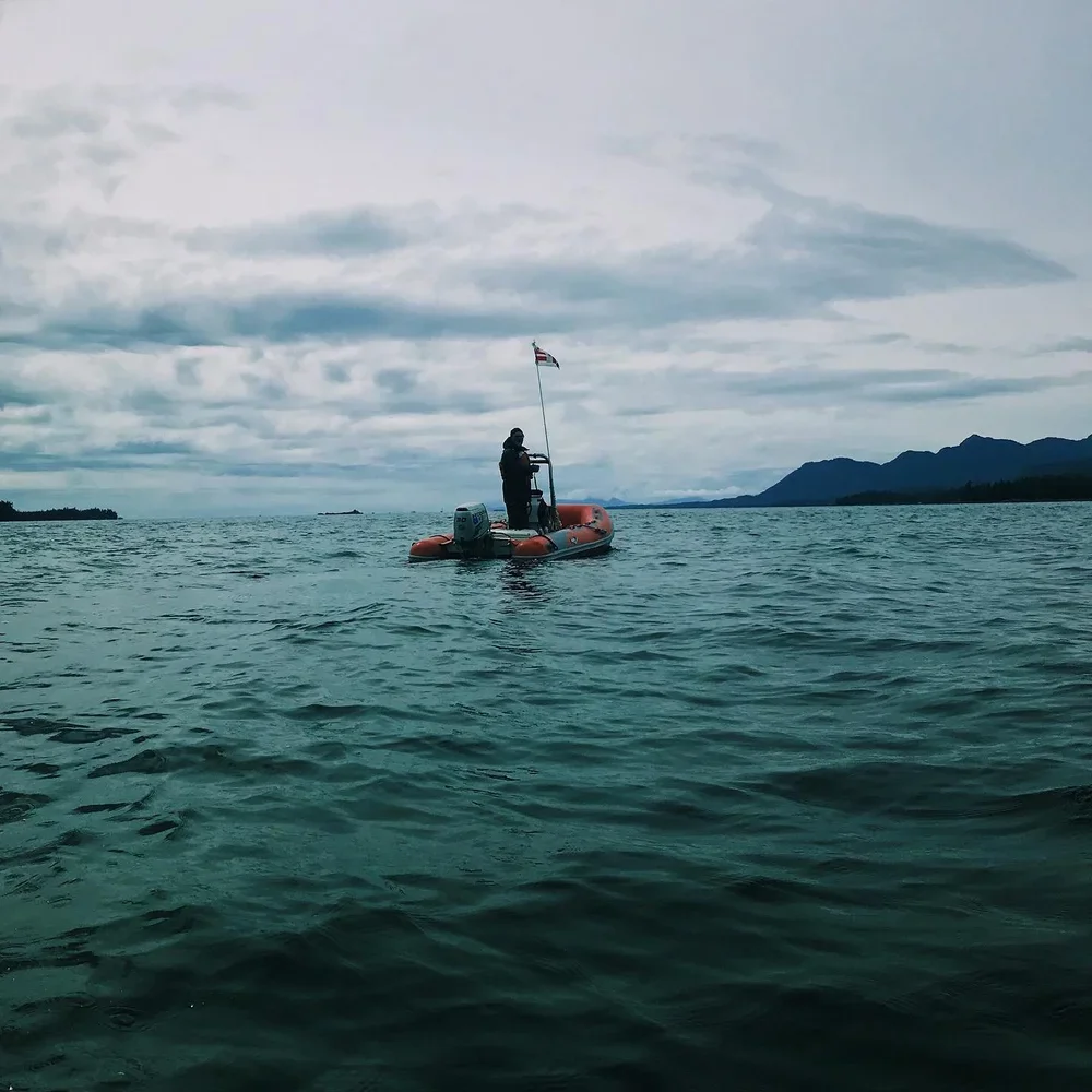 Person standing on a small orange inflatable boat with an outboard motor, holding a fishing rod and a flag, on a calm body of water with mountains in the background.