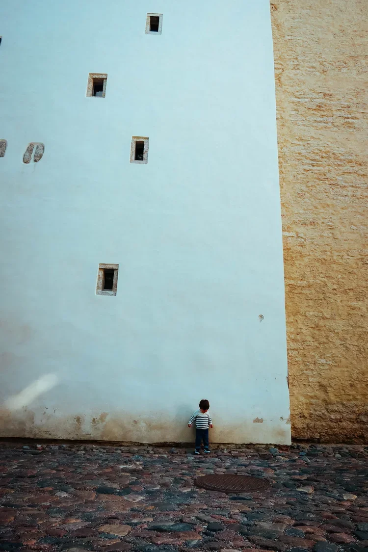 A small child wearing a striped shirt and dark pants standing in front of a large pale yellow wall with small windows, on a cobblestone street.