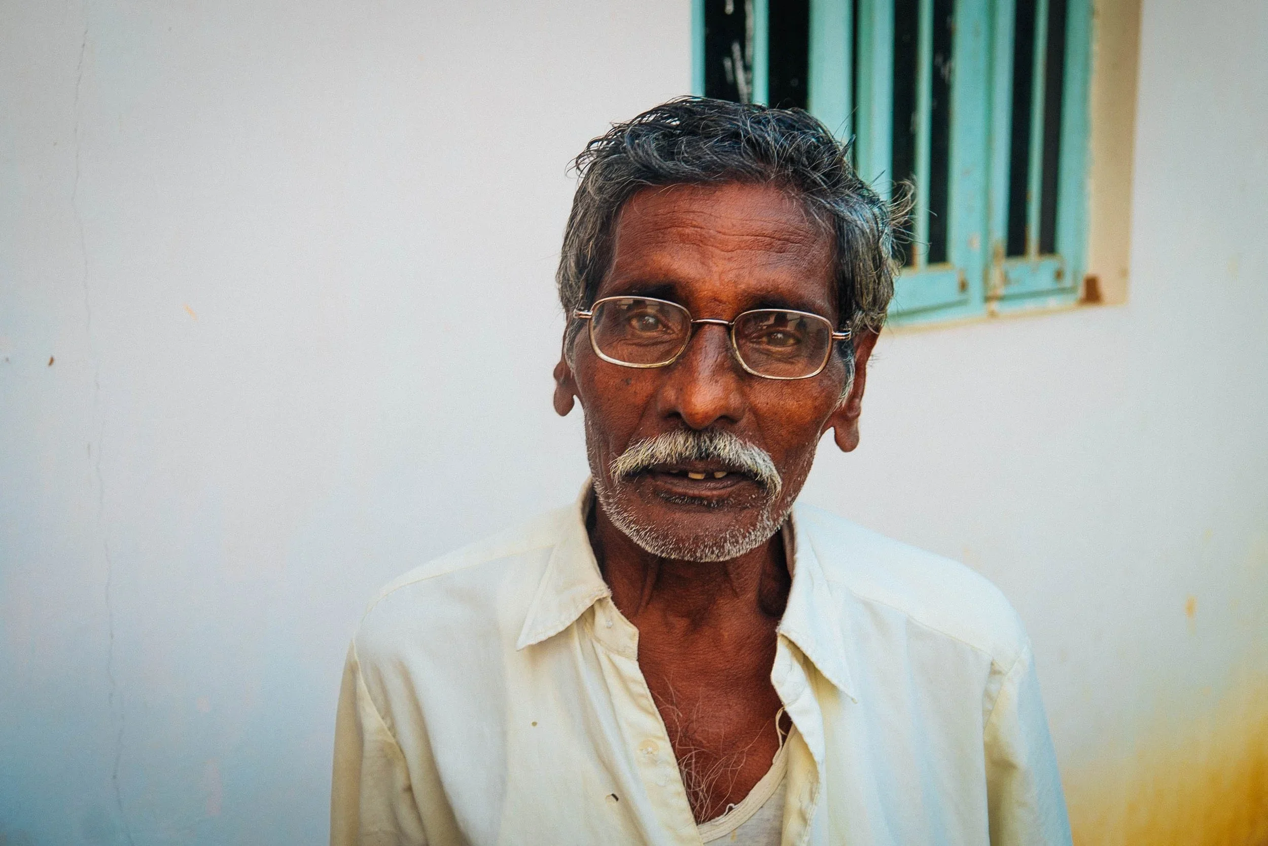 An elderly man with gray hair, a mustache, and glasses, standing against a plain white wall with a small window behind him.