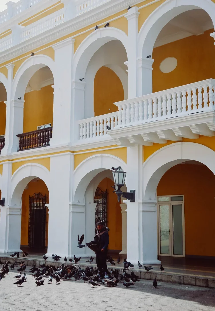 A man standing on a paved sidewalk surrounded by pigeons in front of a yellow building with white arches and columns.