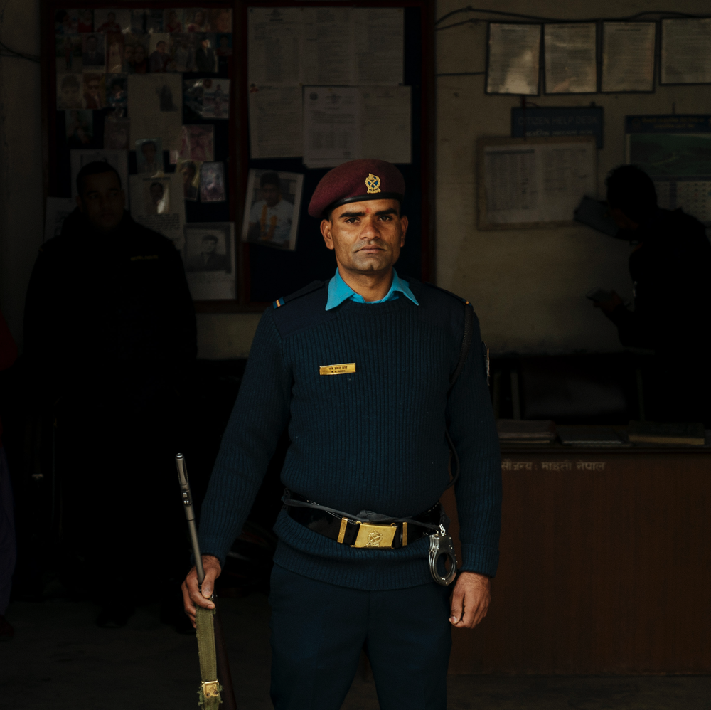 A police officer standing inside a room, holding a baton in his right hand, wearing a maroon beret, and a blue uniform with a badge.