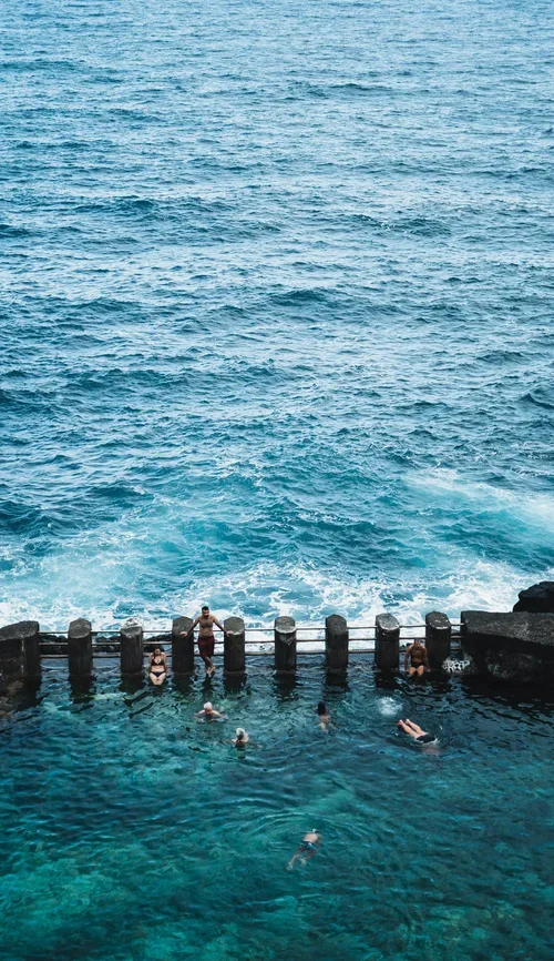 People swimming and relaxing in a pool near the ocean, with waves crashing against the rocks behind.