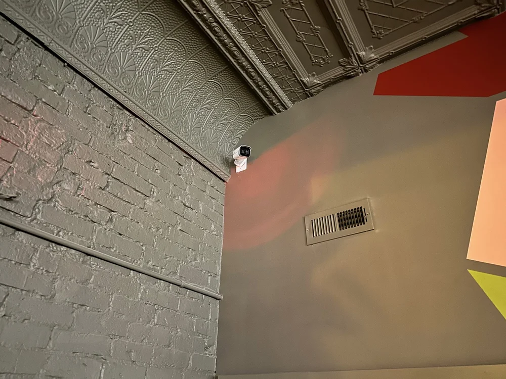 Compact security camera mounted where a brick wall meets a pressed-tin ceiling inside a commercial space