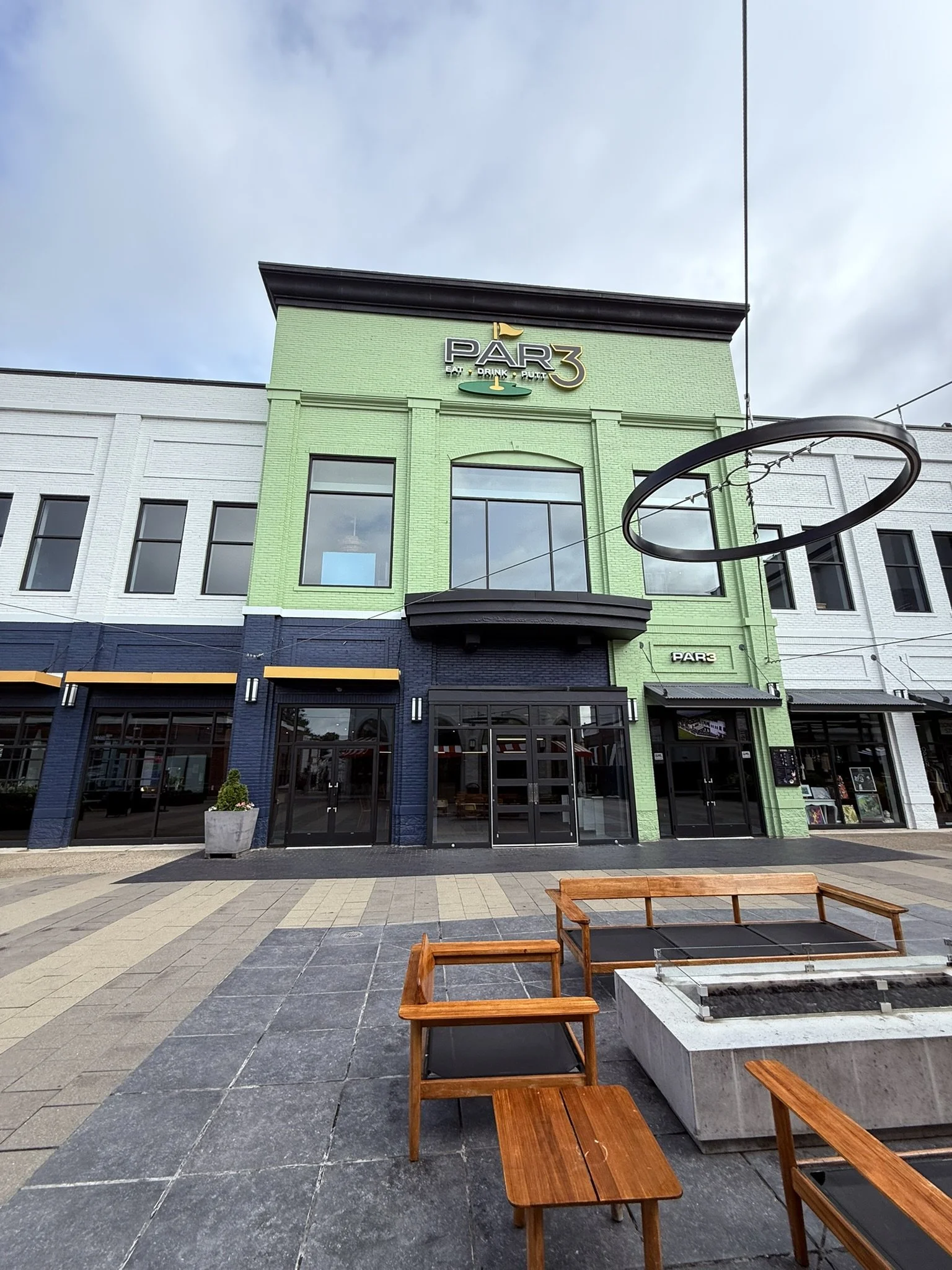 A modern outdoor shopping center with storefronts, benches, and planters, featuring a building with a green and white facade and a sign that says 'PAR3'.