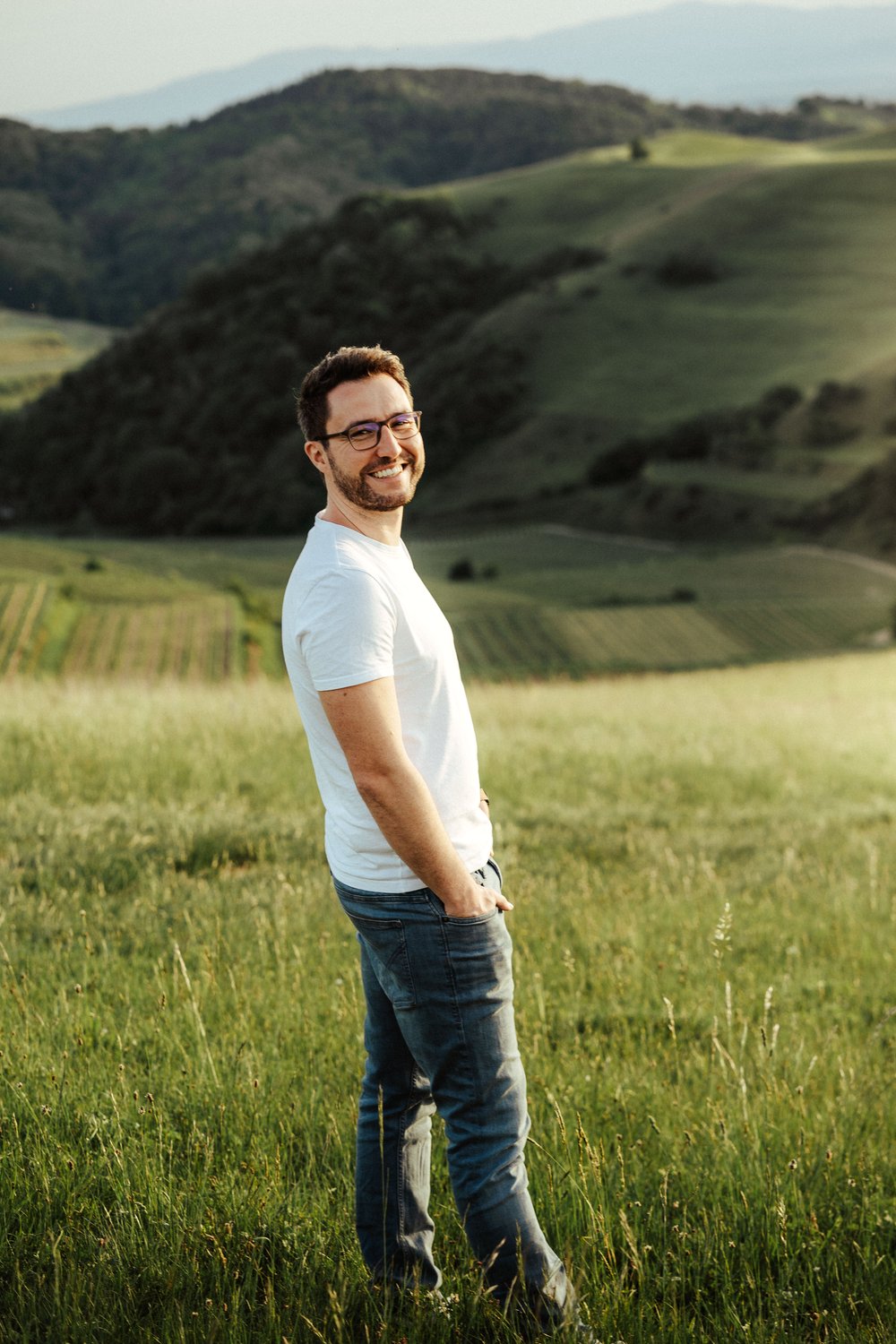 Mann mit Jeans und weißem T-Shirt, Bart, Brille in Abendsonne auf Wiese Blick auf Kaiserstuhl und die Vogesen by UhlArt Fotografie