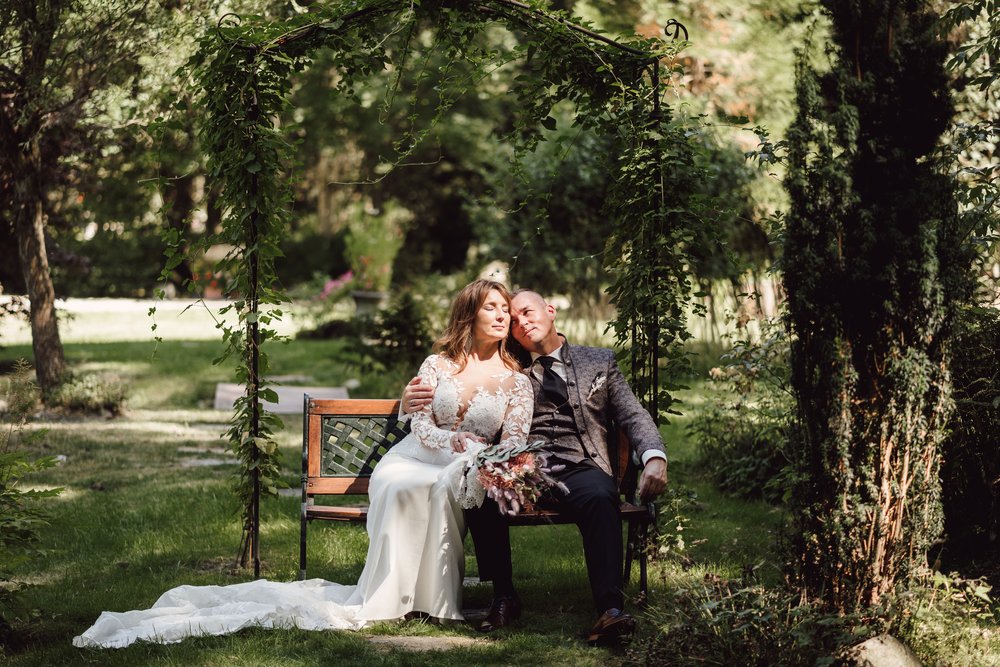 Brautpaar entspannt auf geschwungener Parkbank, Licht fällt  durchs die Bäume